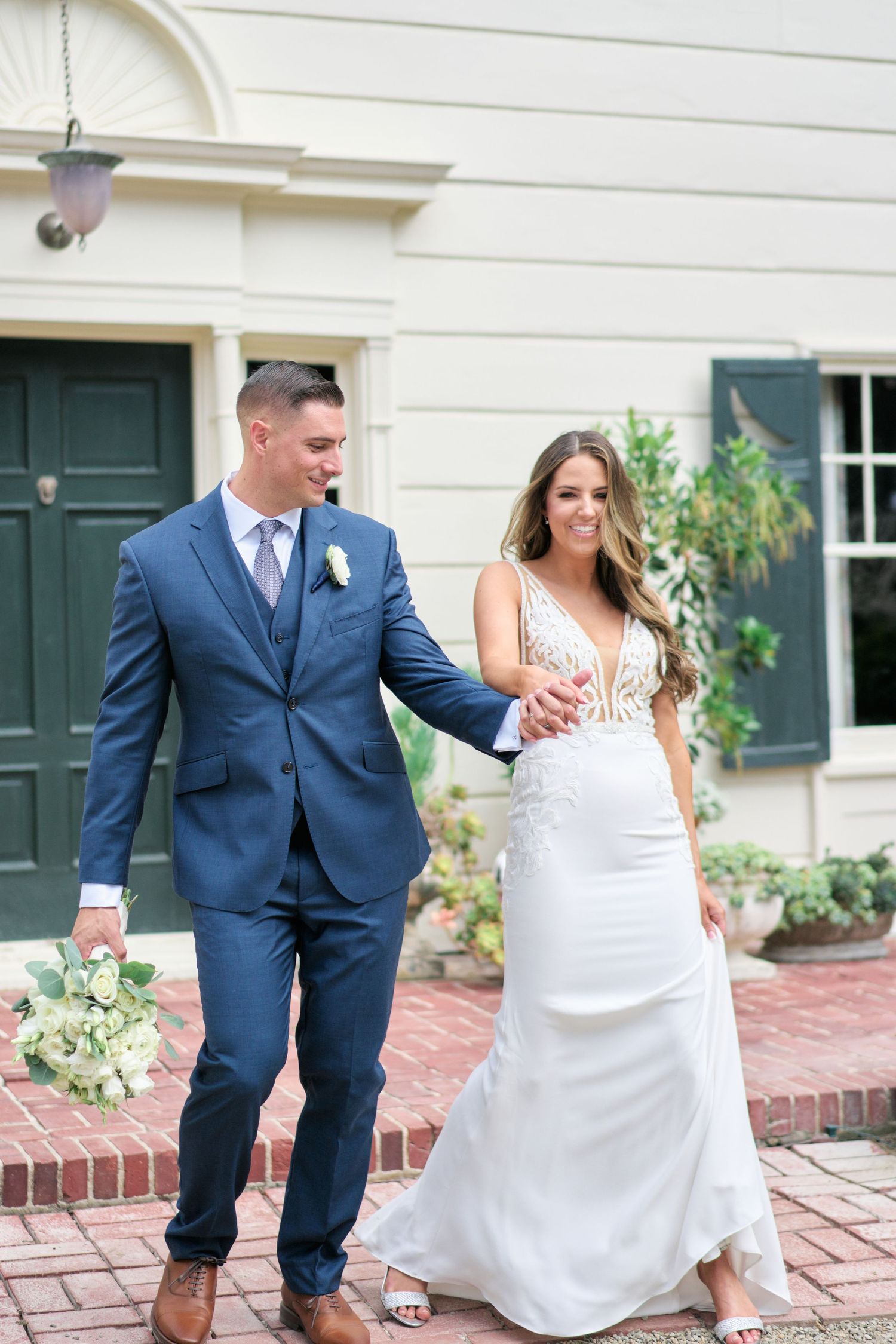 Couple in blue suit and white gown walking together on brick patio in front of historic building.