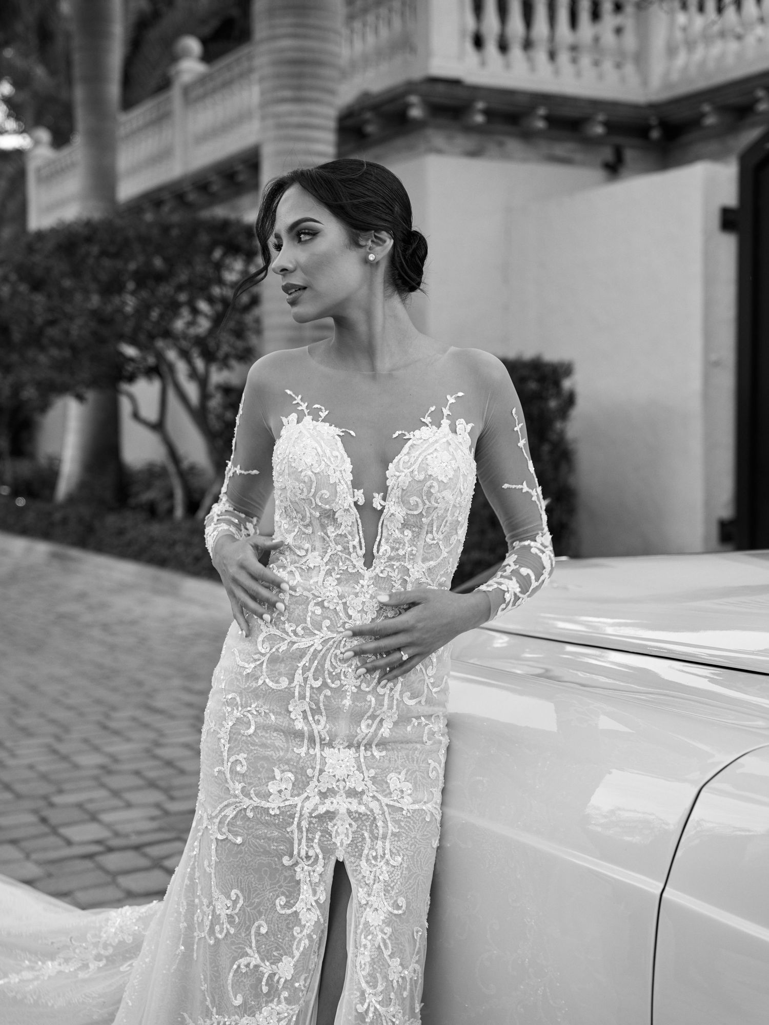 A stunning lace wedding gown with illusion sleeves and plunging neckline shown outdoors against a white building.
