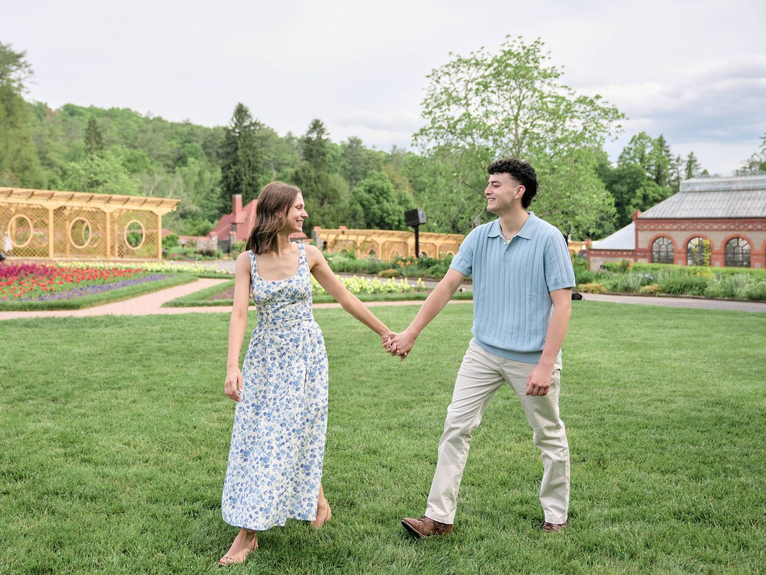 A couple holds hands and smiles while walking through a lush green garden with colorful flowers and trees.
