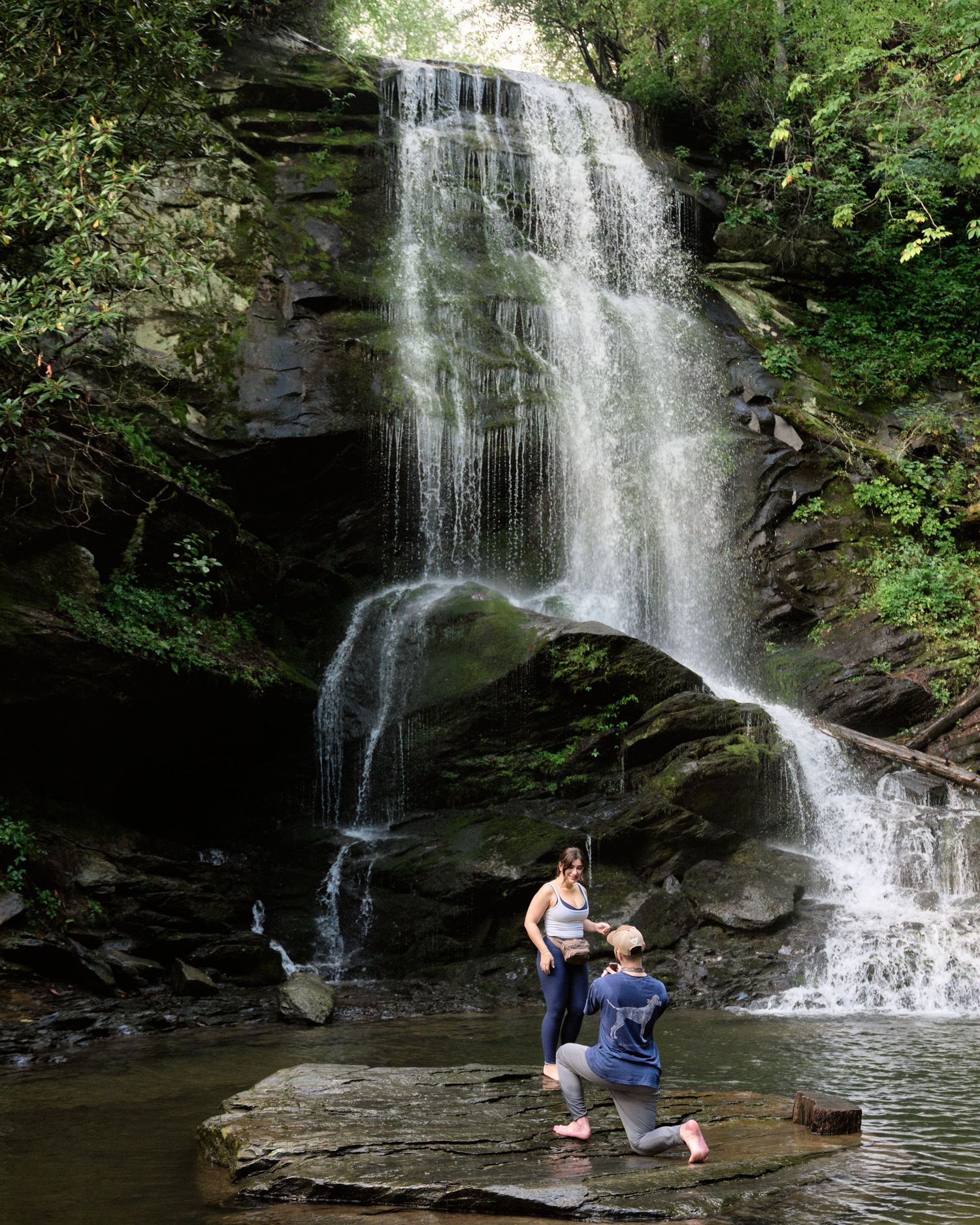 Person proposing on a rock ledge in front of a stunning cascading waterfall surrounded by lush green forest.