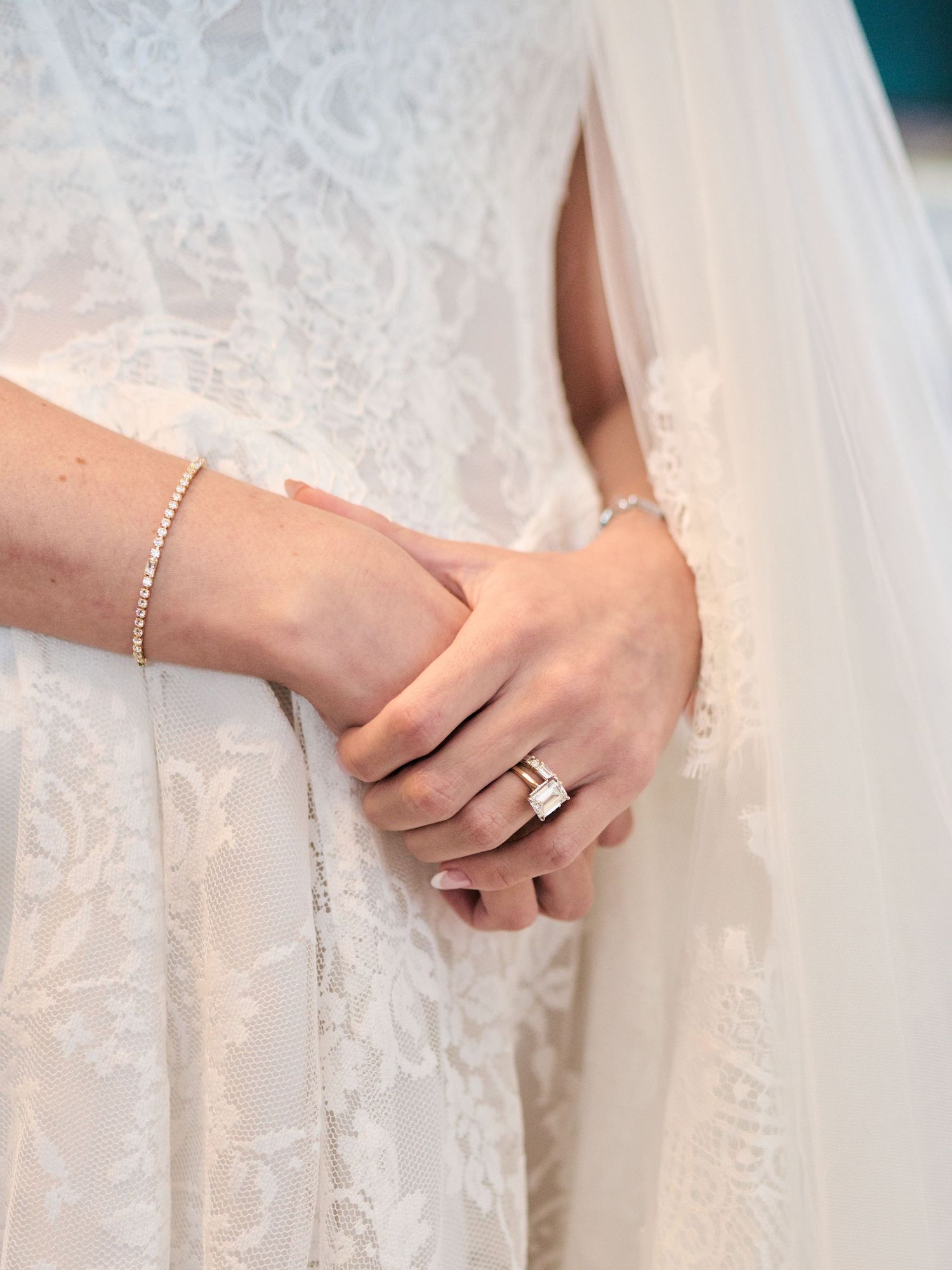 Close-up of bride's hands clasped together wearing a diamond engagement ring, wedding band, and delicate bracelet.
