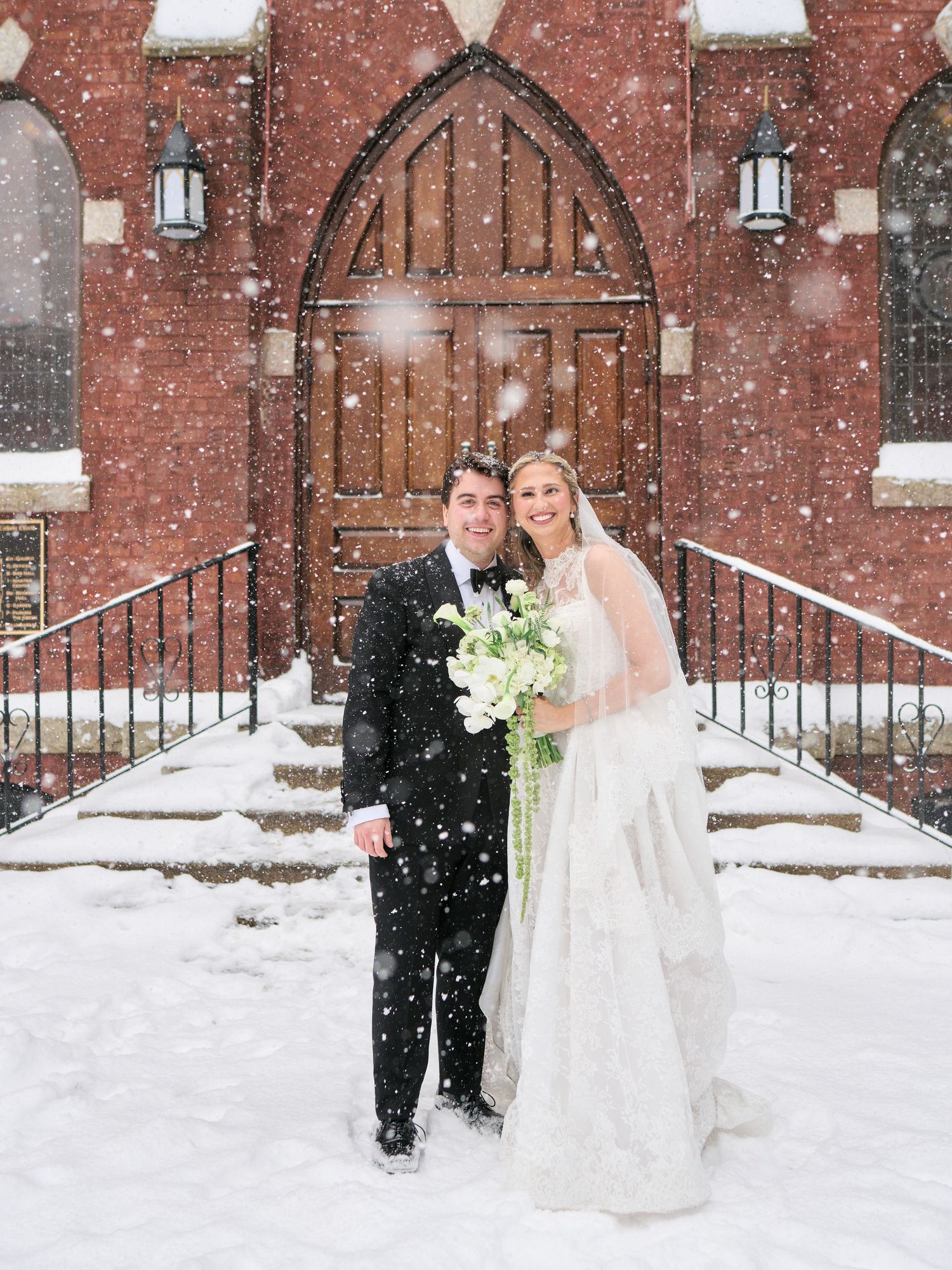 Bride and groom pose in falling snow outside a brick church with gothic arch doors, bride holding white bouquet.