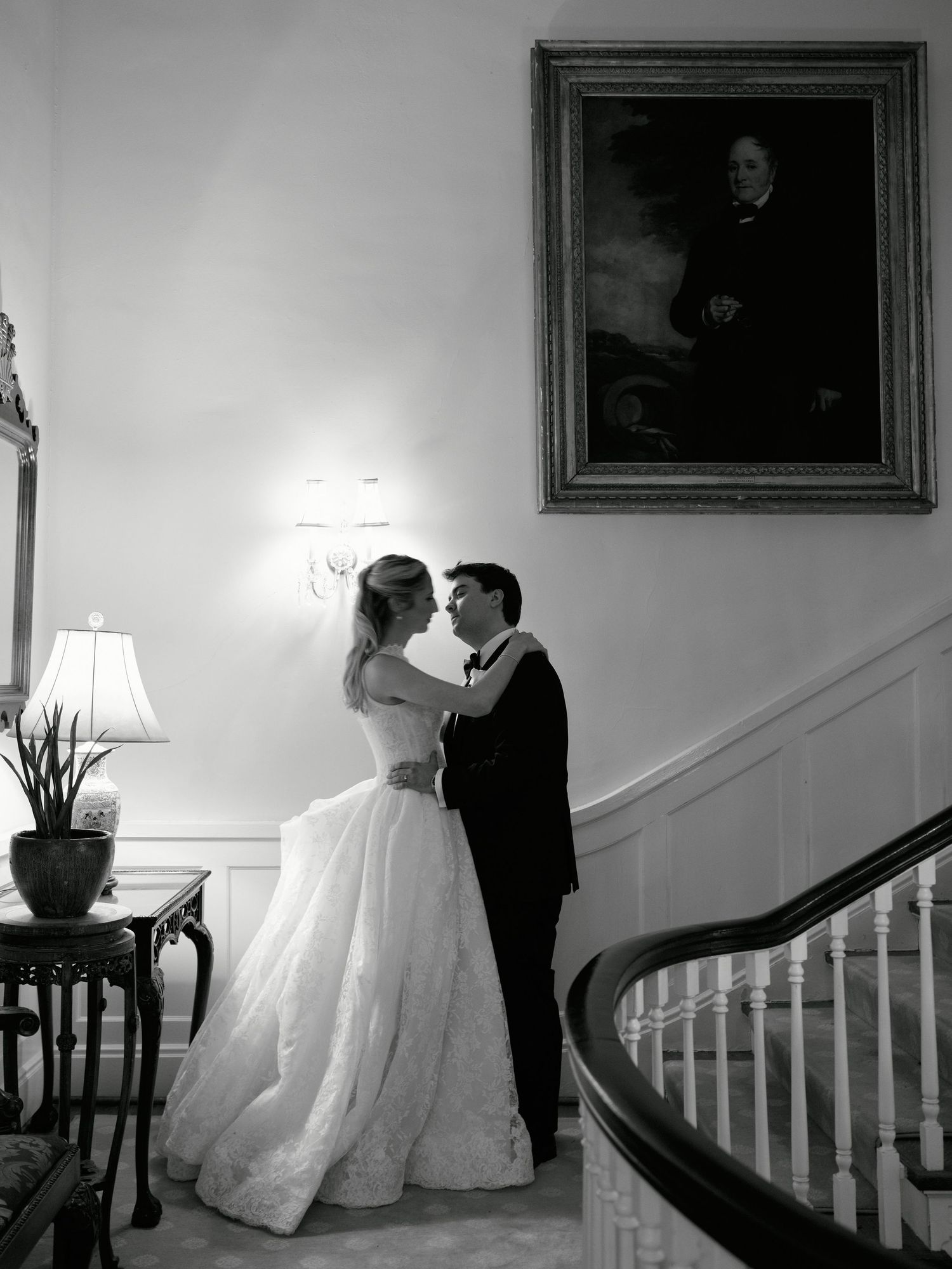 Black and white photo of a bride and groom sharing a romantic moment by a staircase beneath a portrait painting.