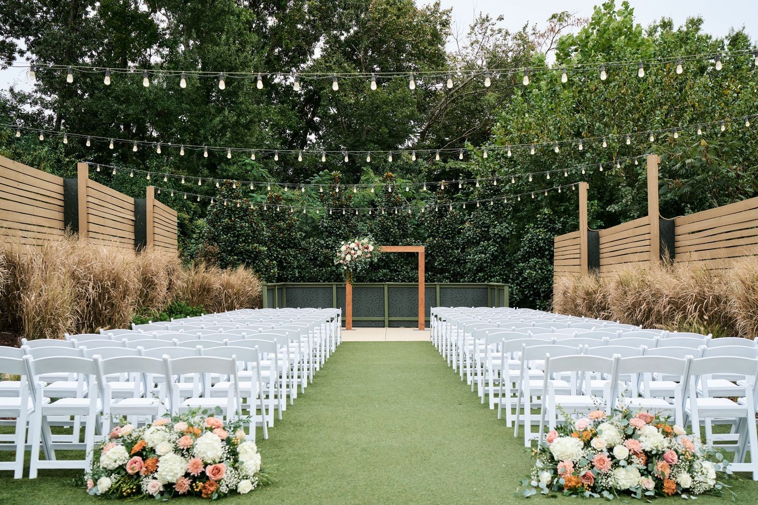 Elegant outdoor wedding ceremony setup with white chairs, string lights, floral arrangements, and a wooden arch on lush green grass.