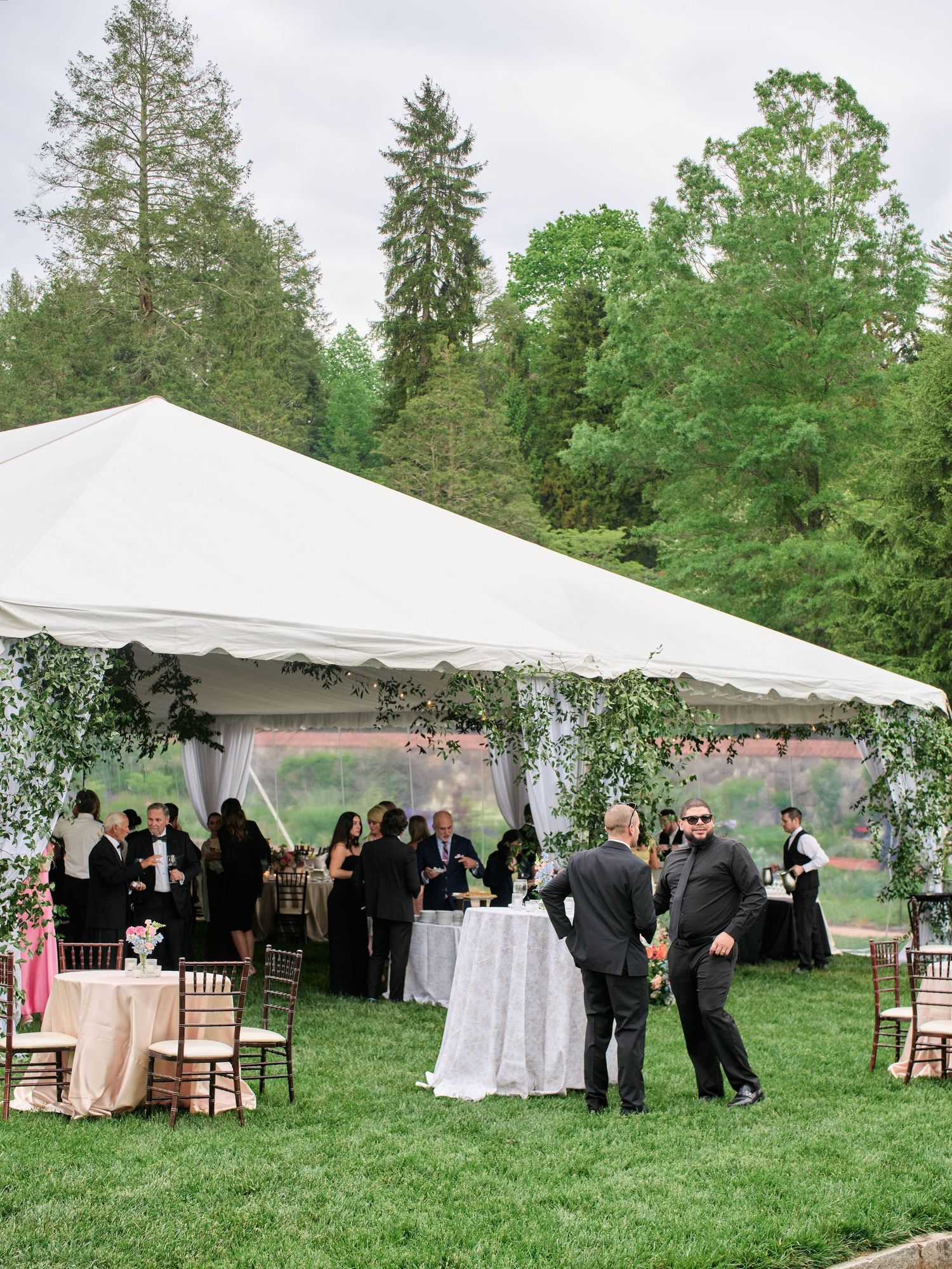 Outdoor wedding reception under a white tent with tables and chairs set up on a green lawn surrounded by tall evergreen trees.