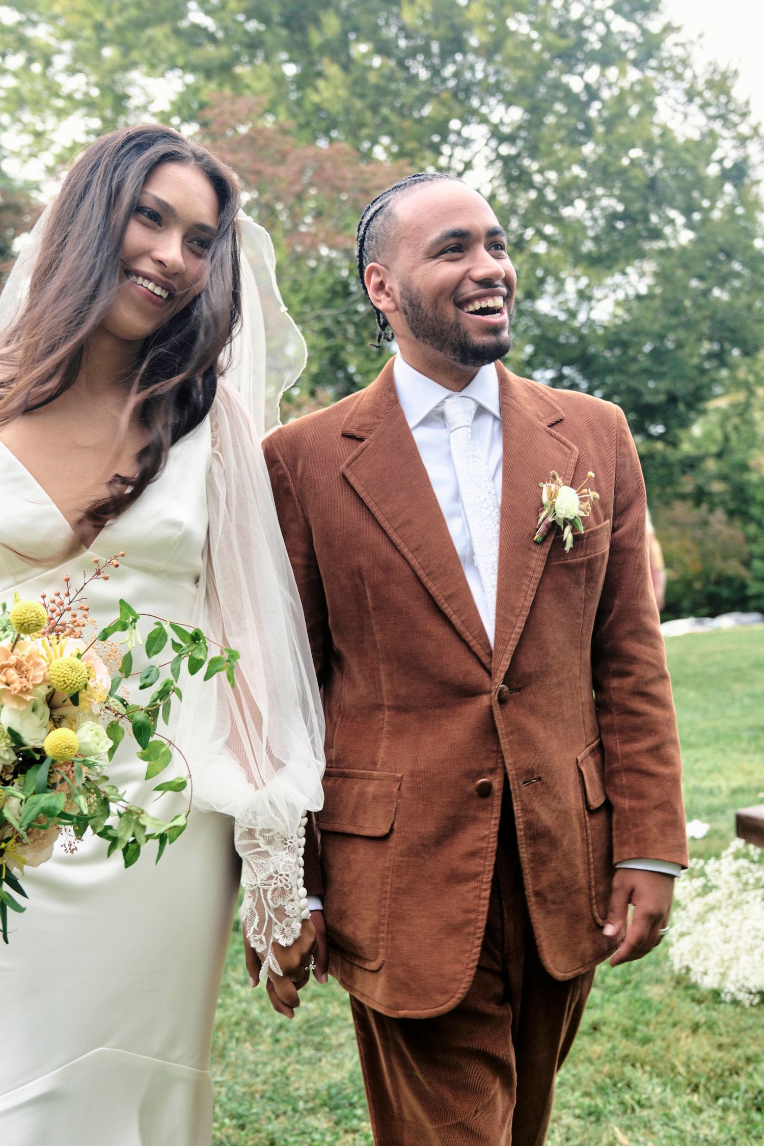 A bride in a white gown holds a yellow bouquet while standing with her groom in a brown suit at their outdoor wedding.