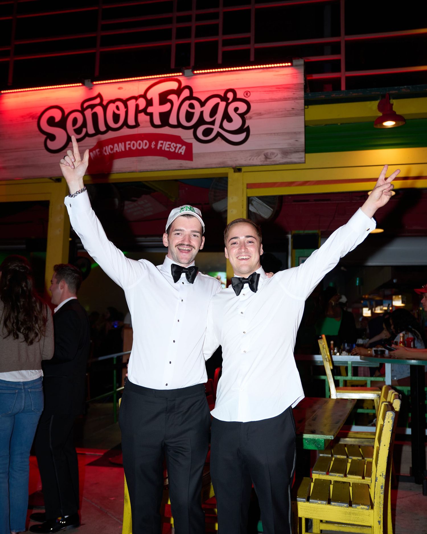 Two waiters in white dress shirts and bow ties celebrate with raised arms in front of a Senor Frog's restaurant sign.