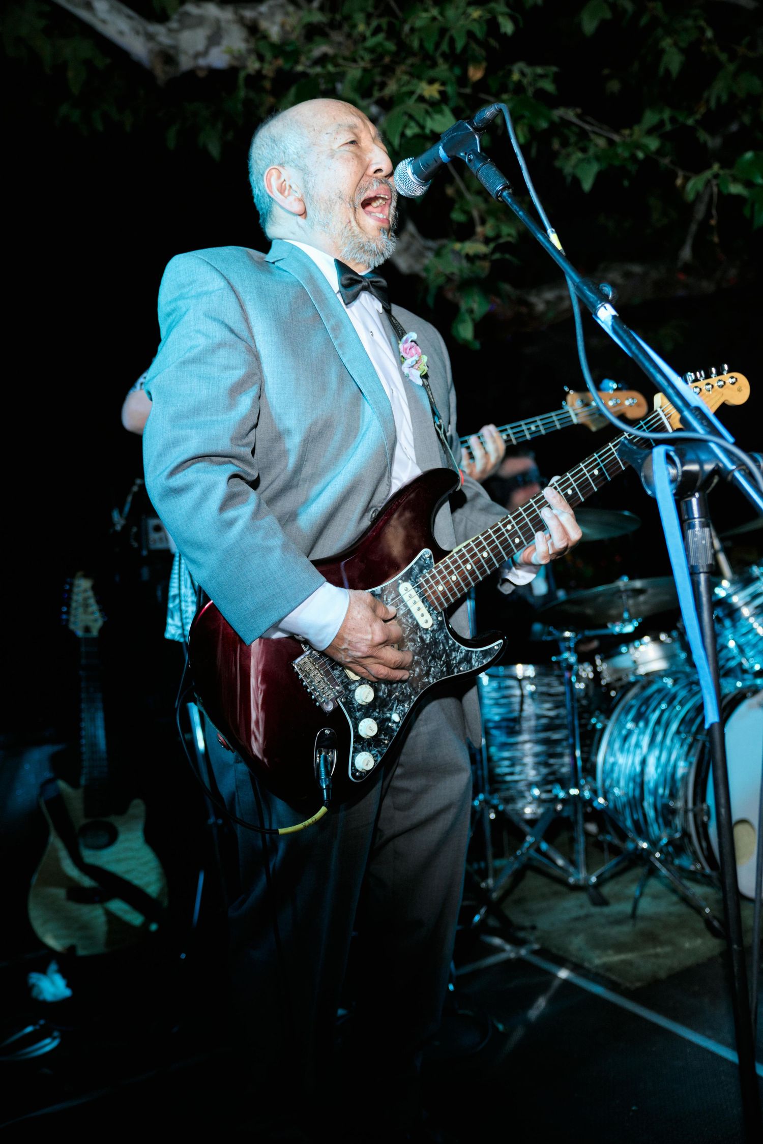 Musician in grey suit playing electric guitar and singing at outdoor evening event.