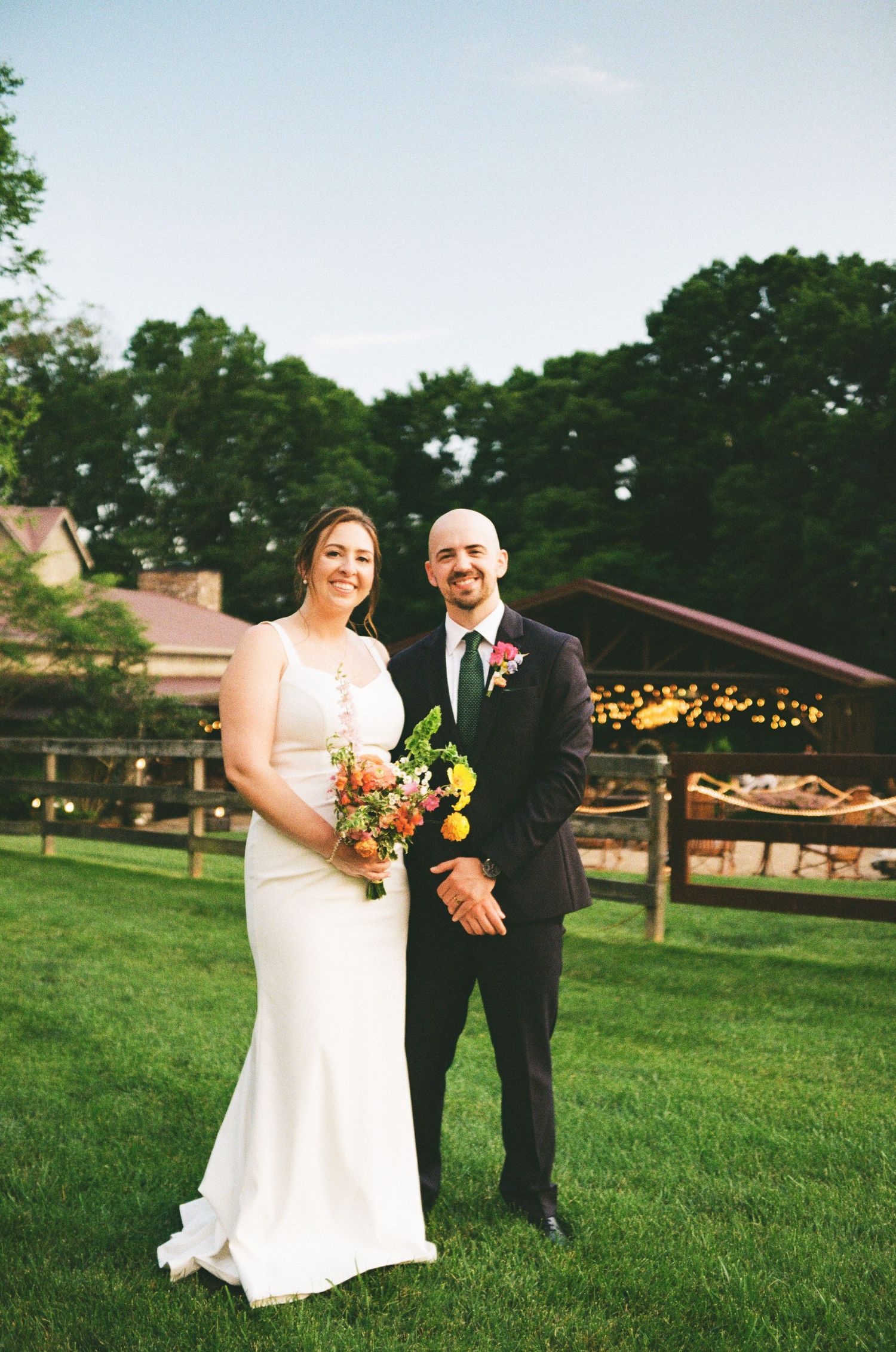 A wedding portrait taken on a grassy lawn with rustic farm buildings and fences in the background.