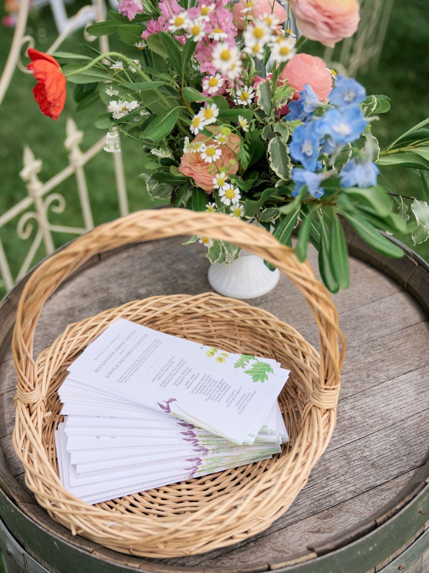 Floral arrangement with pink and blue flowers in a vase next to program papers in a woven basket on wooden table.