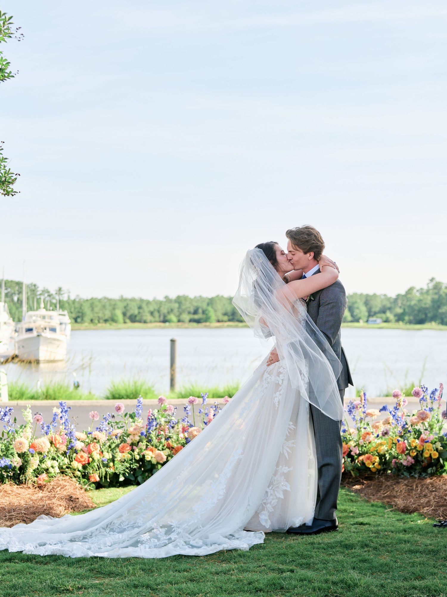 A couple embraces by a lakeside garden with colorful flowers and a flowing white wedding dress on a sunny day.