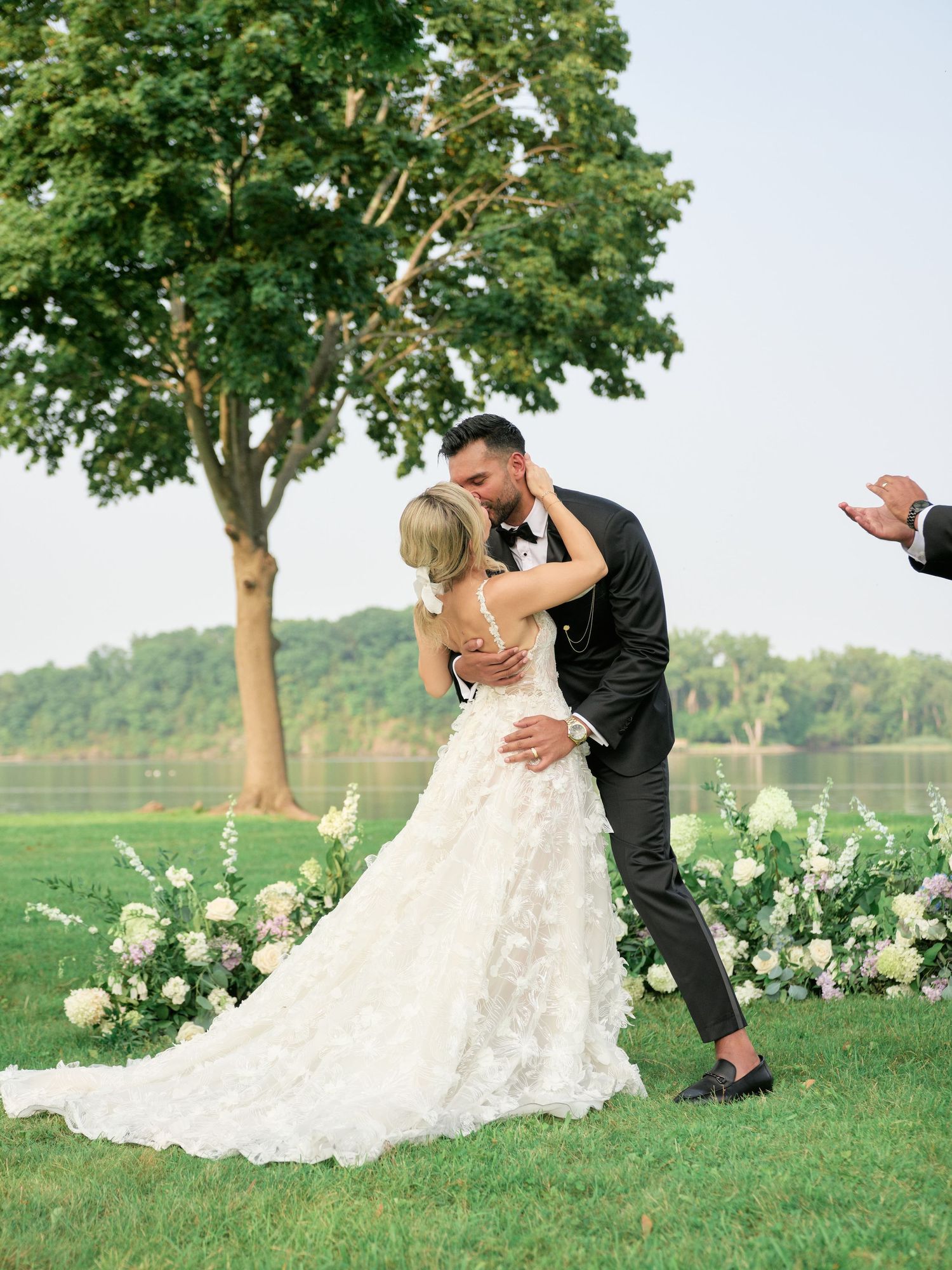 A romantic wedding kiss on a green lawn with white floral arrangements and a tree in the background.