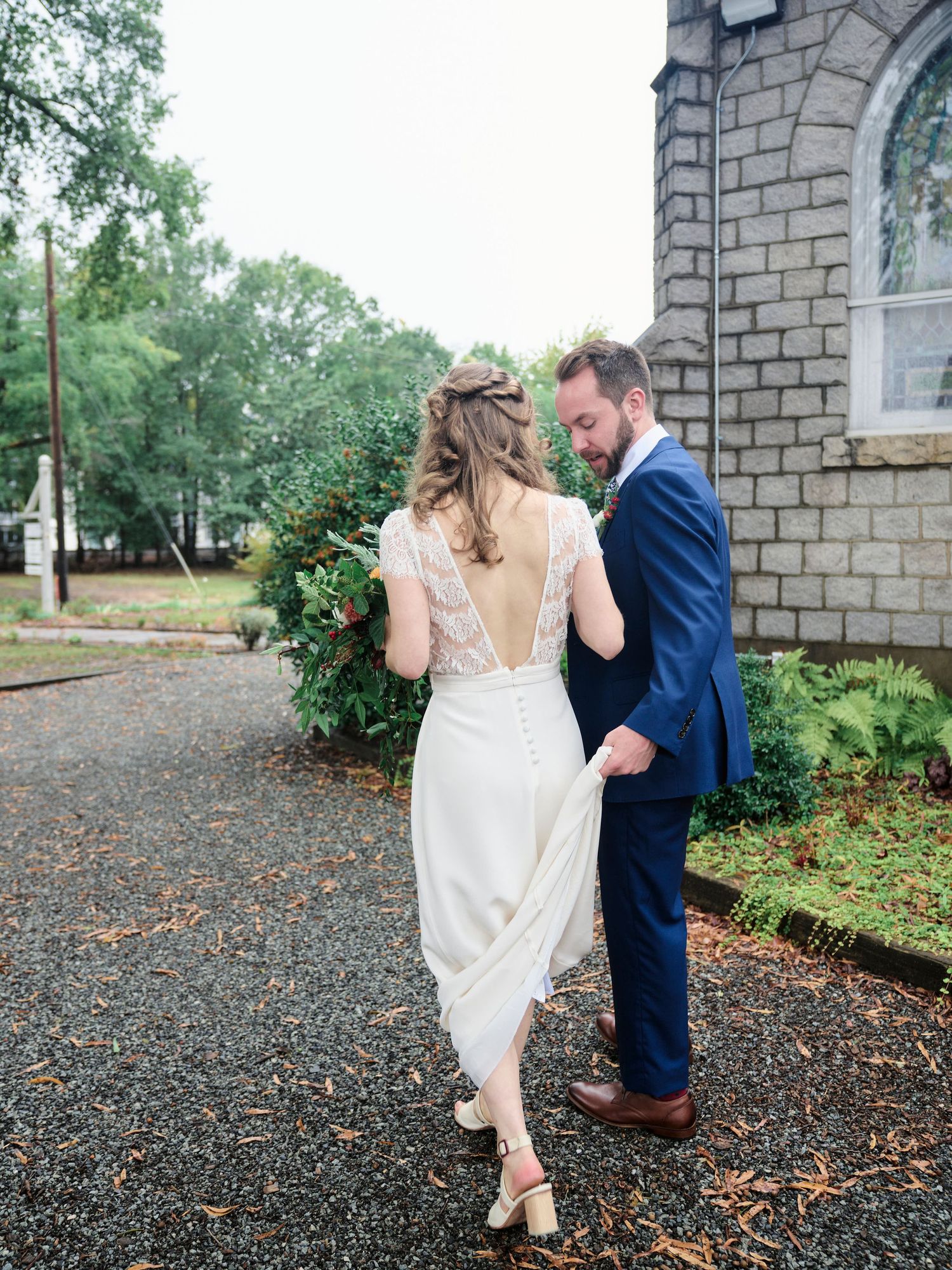 A couple in formal attire walks along a path beside a historic stone church building.
