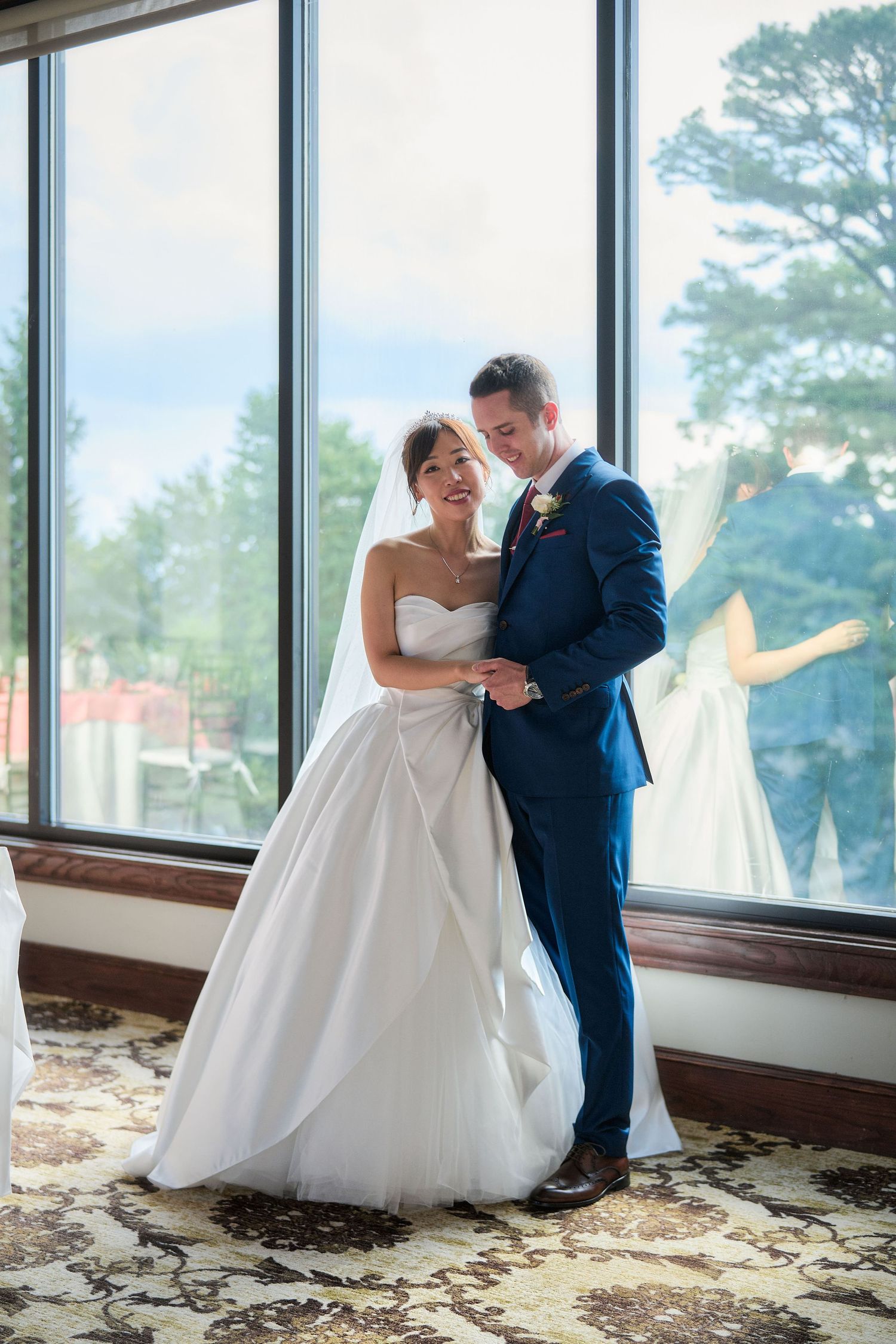 A wedding couple shares a romantic moment beside floor-to-ceiling windows with trees visible outside.