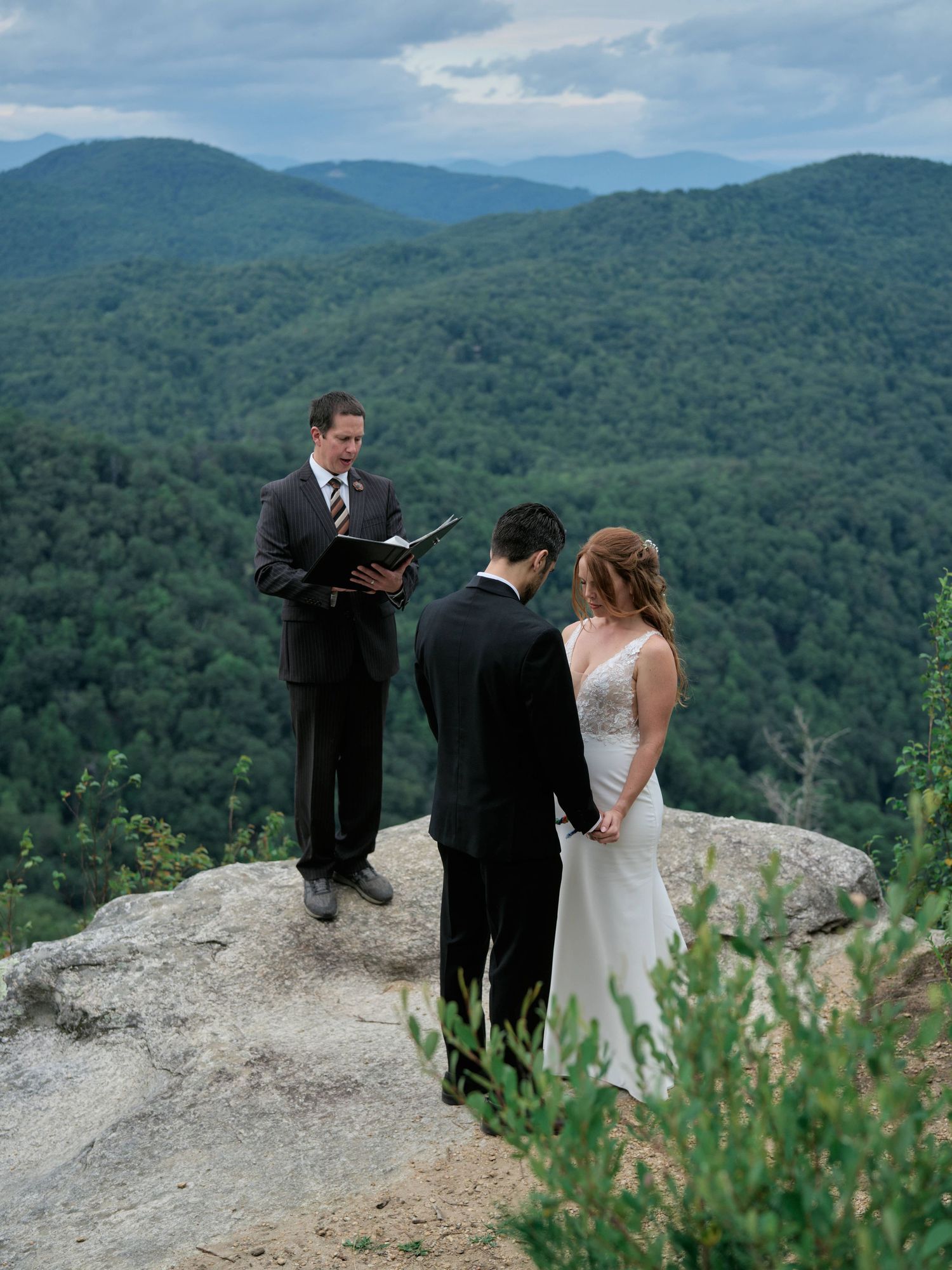 A wedding ceremony takes place on a scenic mountain overlook with rolling green hills stretching into the distance.