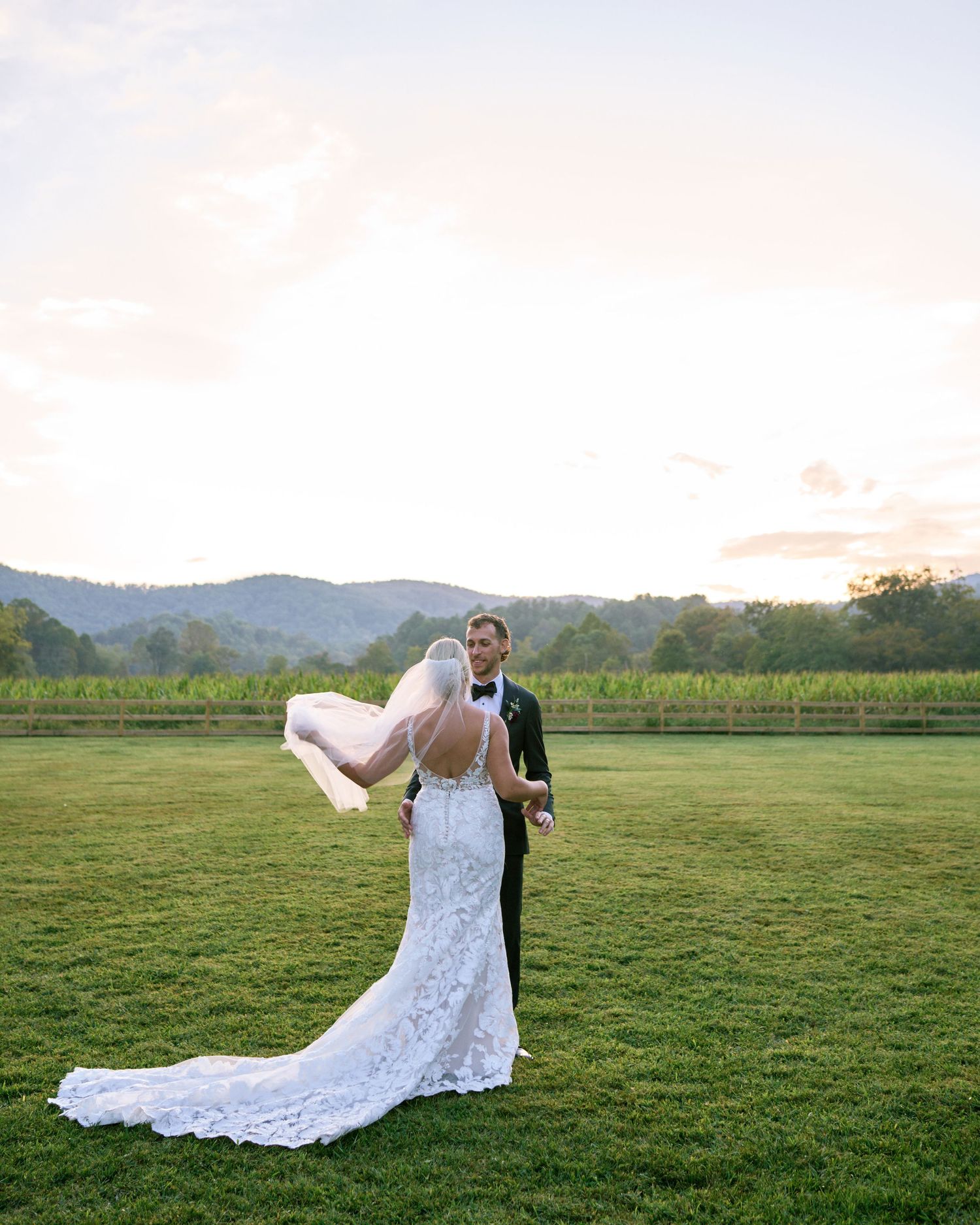 A bride and groom share a romantic moment at sunset on a grassy lawn with mountains and vineyards in the background.
