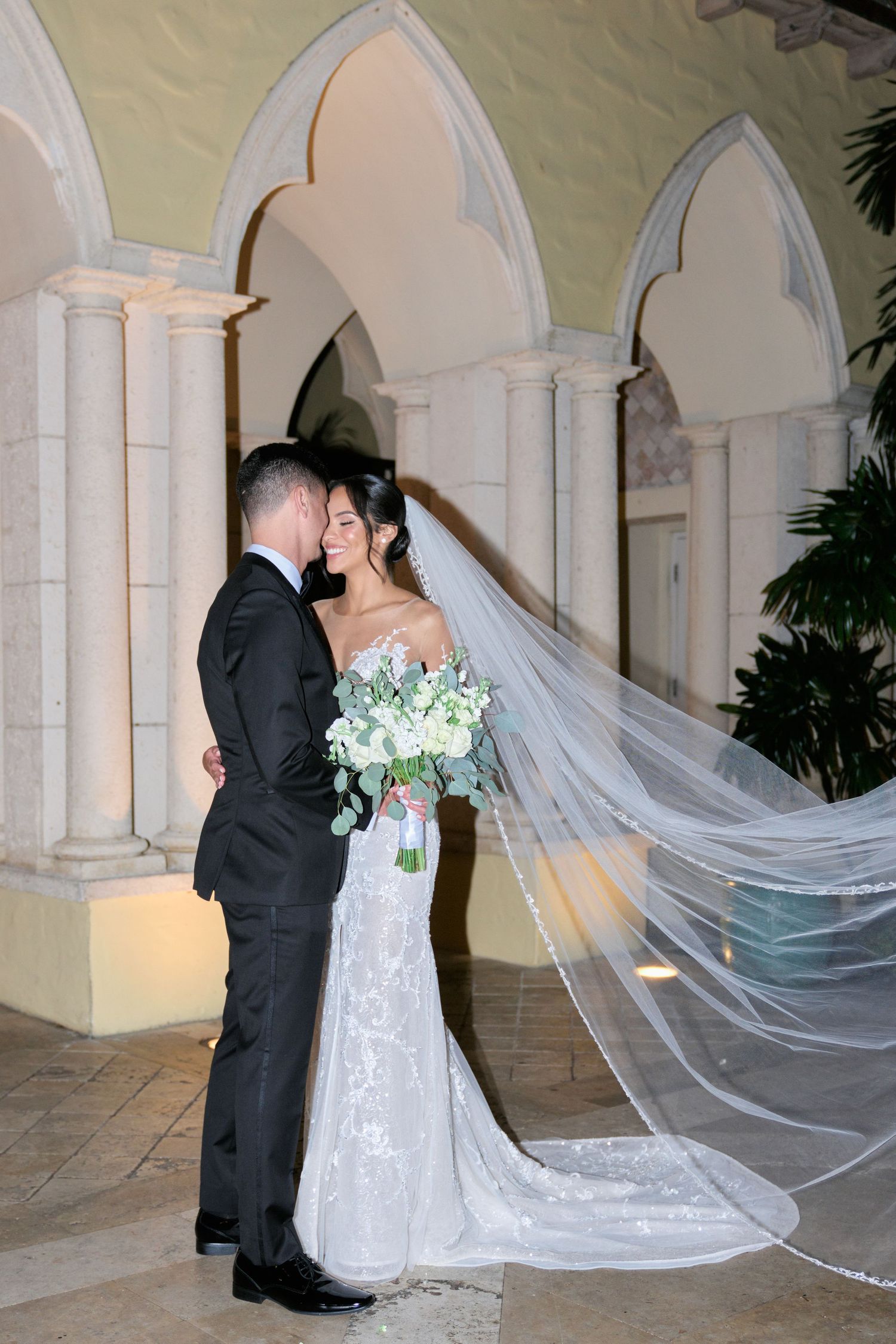 Wedding couple sharing romantic moment under ornate archways with flowing veil and floral bouquet.