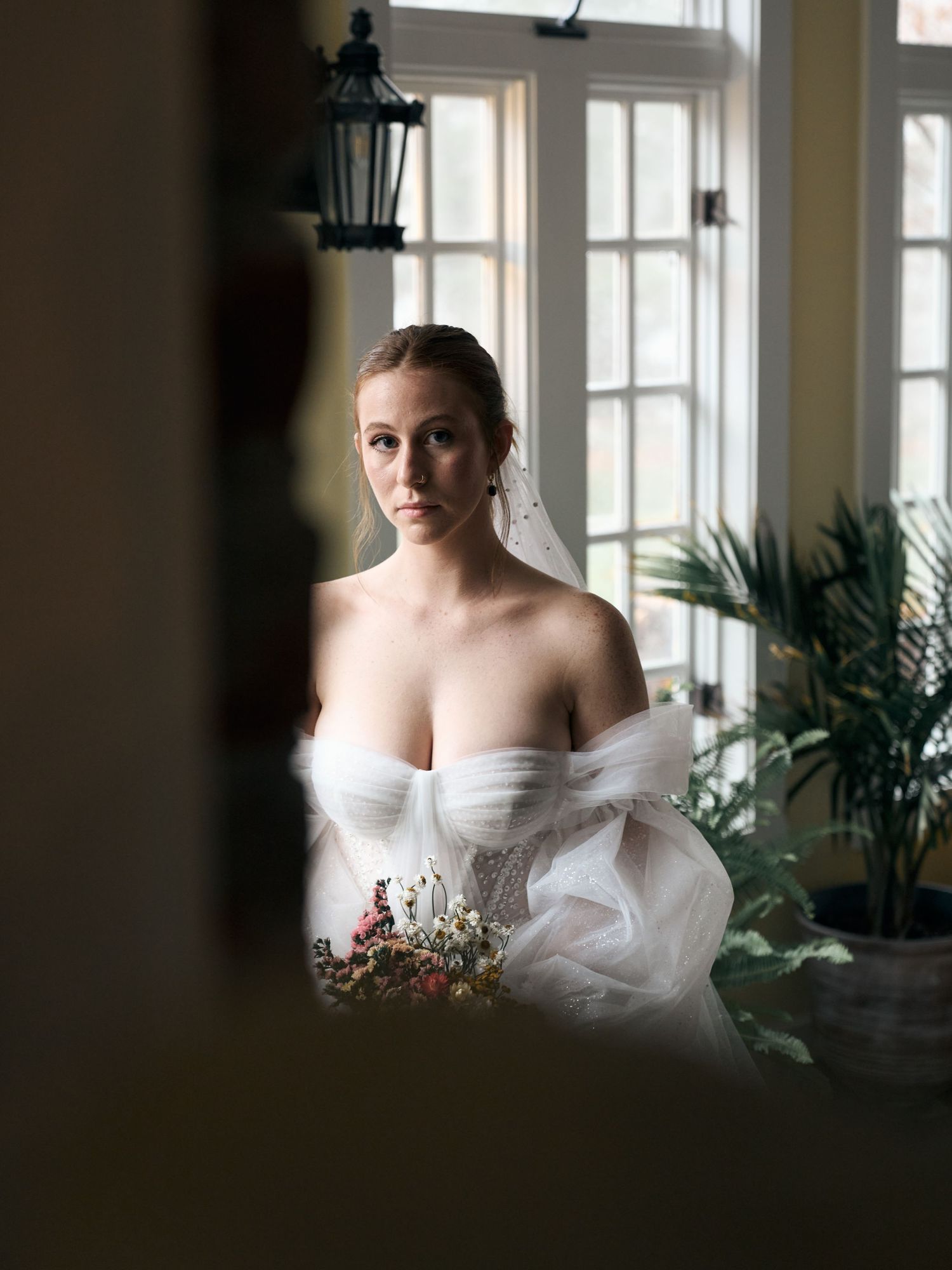 Elegant off-shoulder wedding dress portrait near sunlit windows with delicate florals.