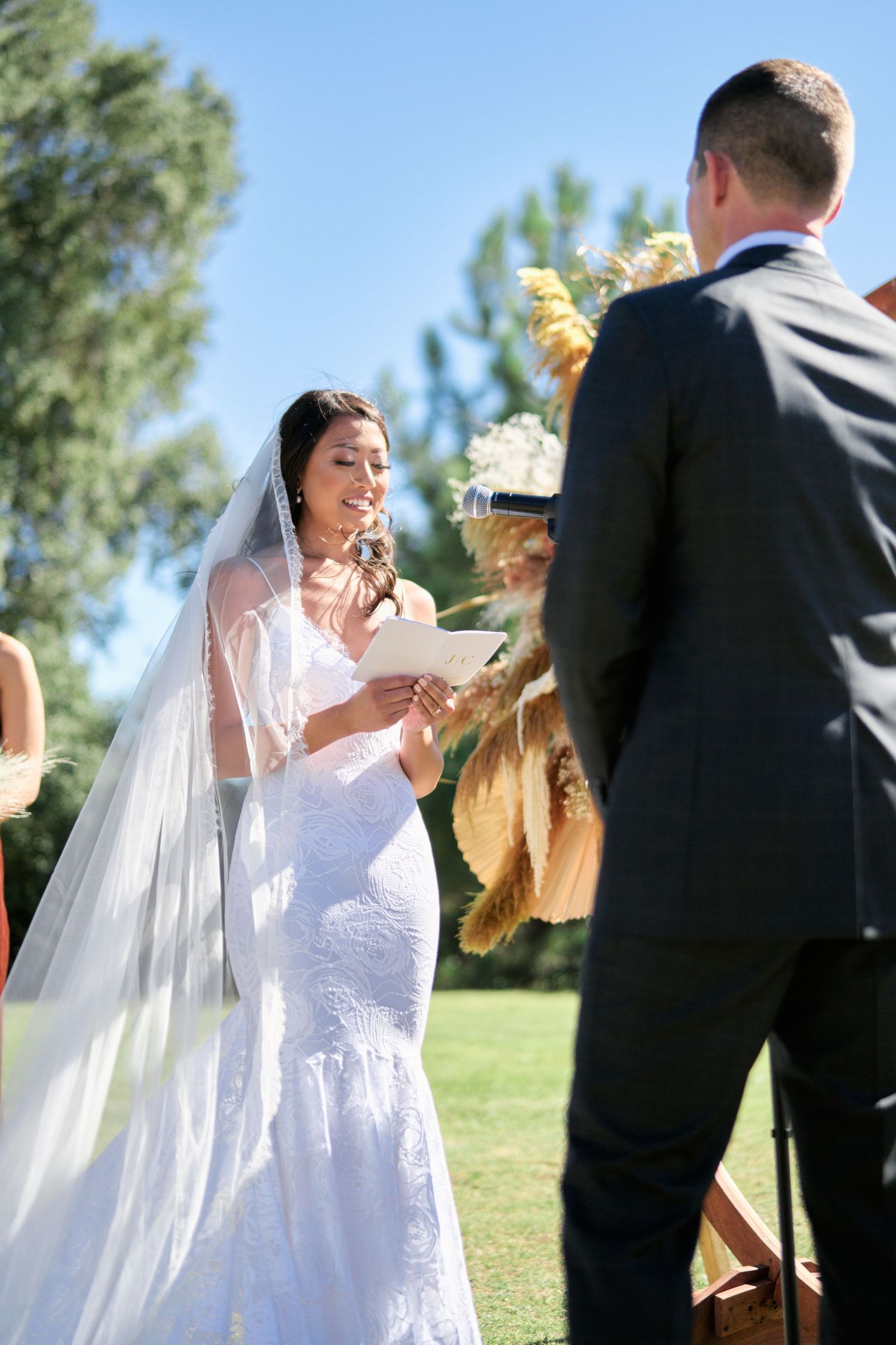 A bride in lace gown reads wedding vows during an outdoor ceremony.