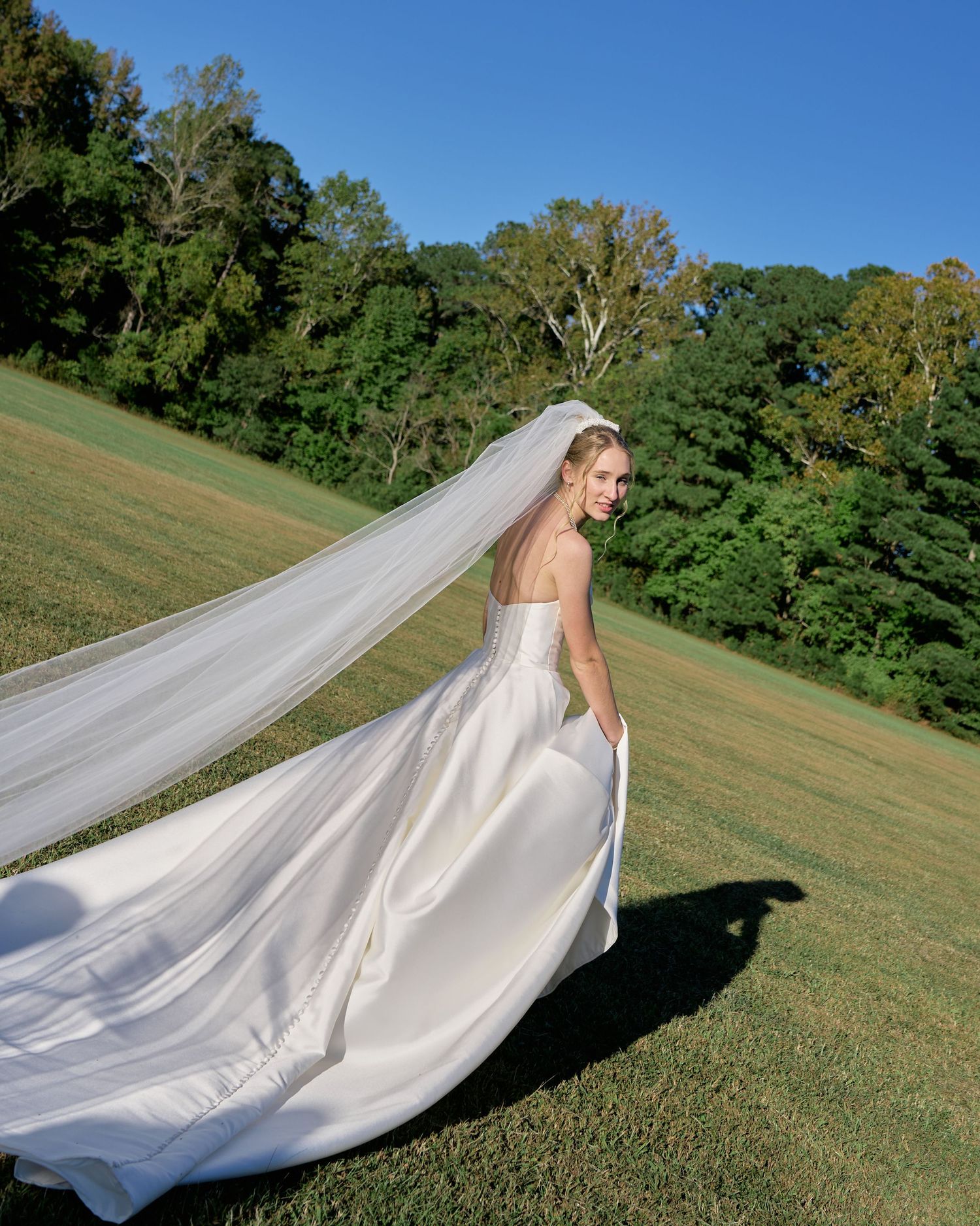 A flowing white wedding veil catches the breeze as it trails dramatically across a grassy hillside on a sunny day.