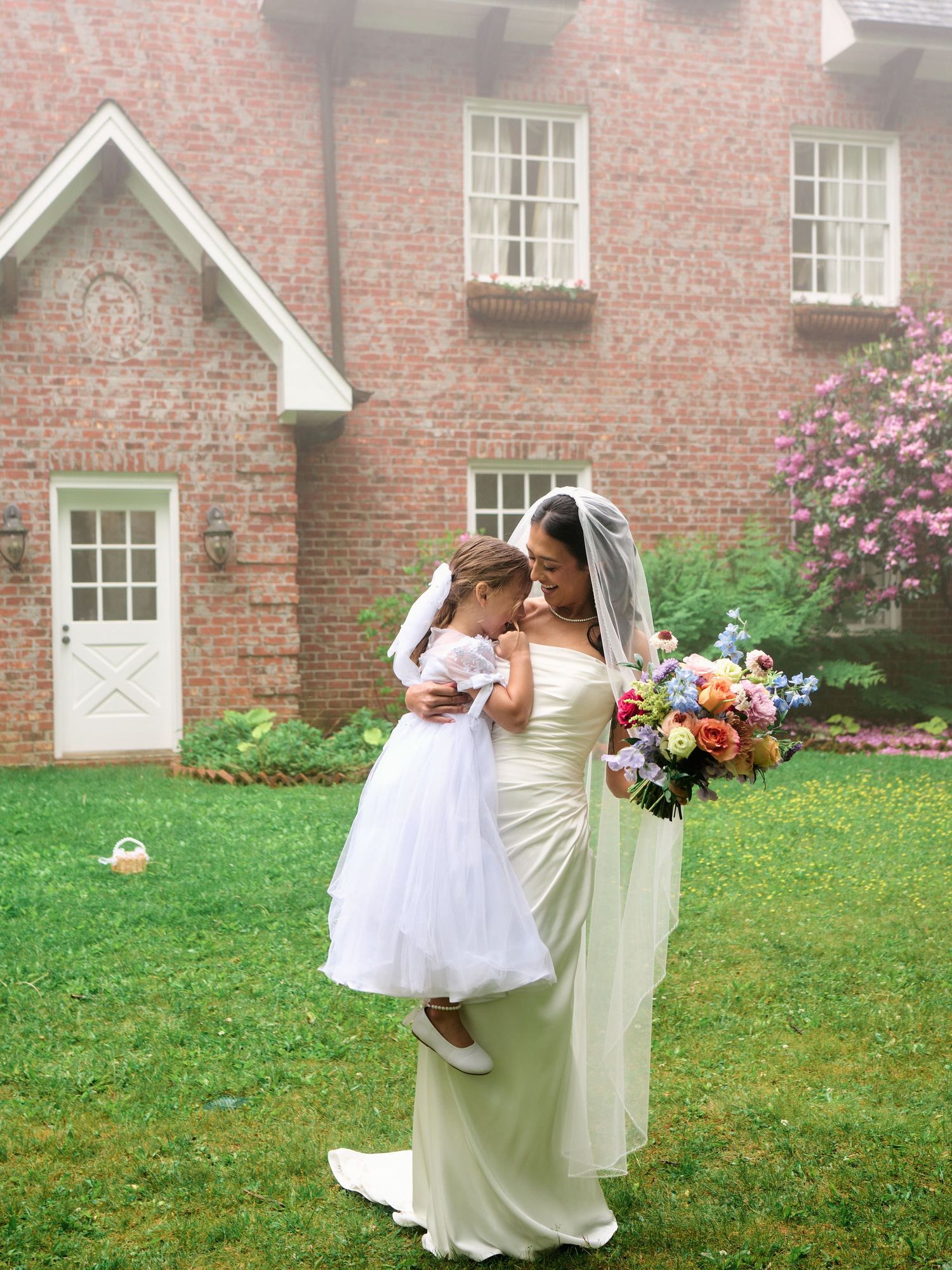 Bride in ivory gown holding flower girl in white dress, posing with colorful bouquet outside brick house.