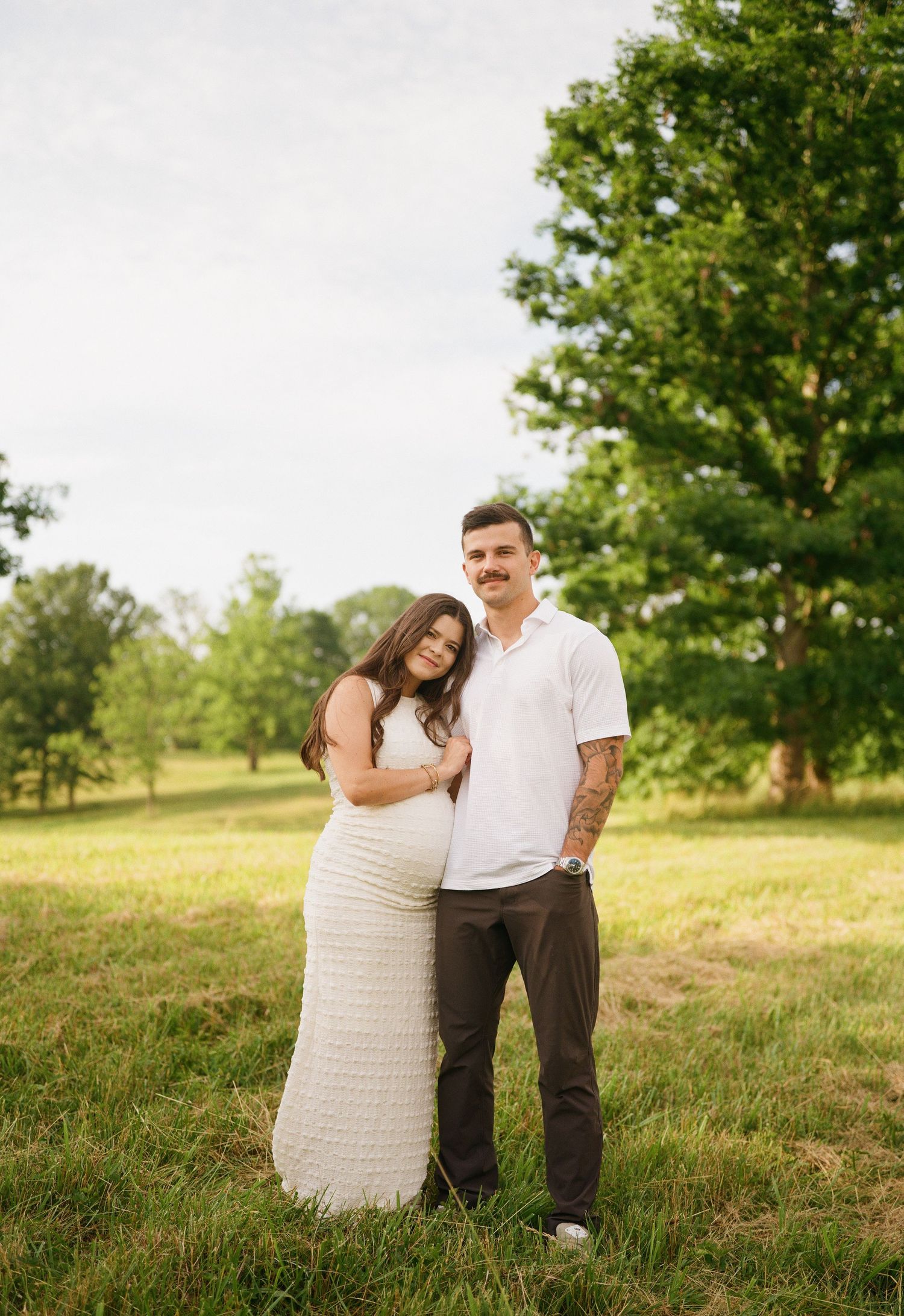 Pregnant couple posing in a green field, woman in white maxi dress leaning on man in white polo and brown pants.