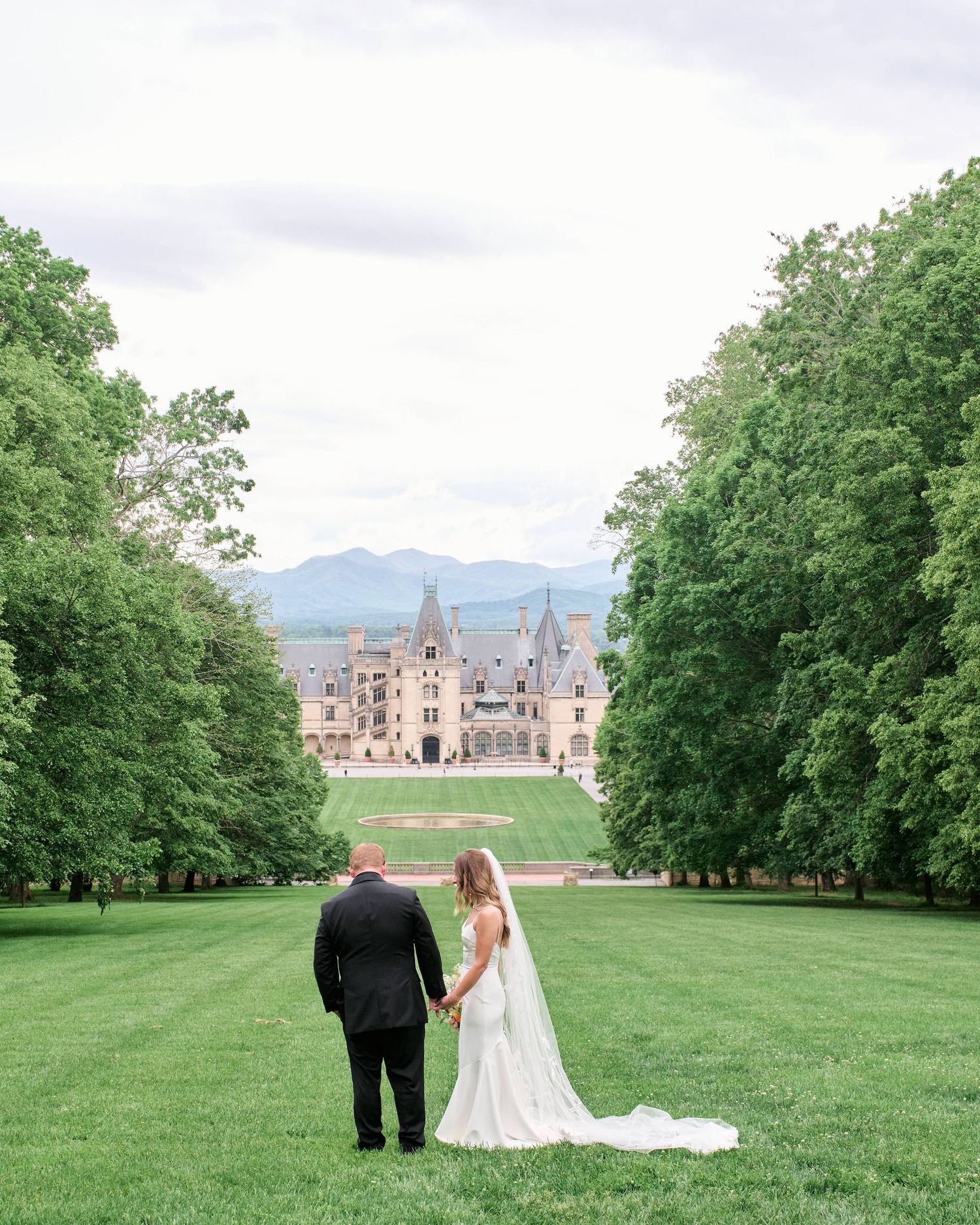 A couple stands on a manicured lawn overlooking the grand Biltmore Estate with mountains visible in the distance.