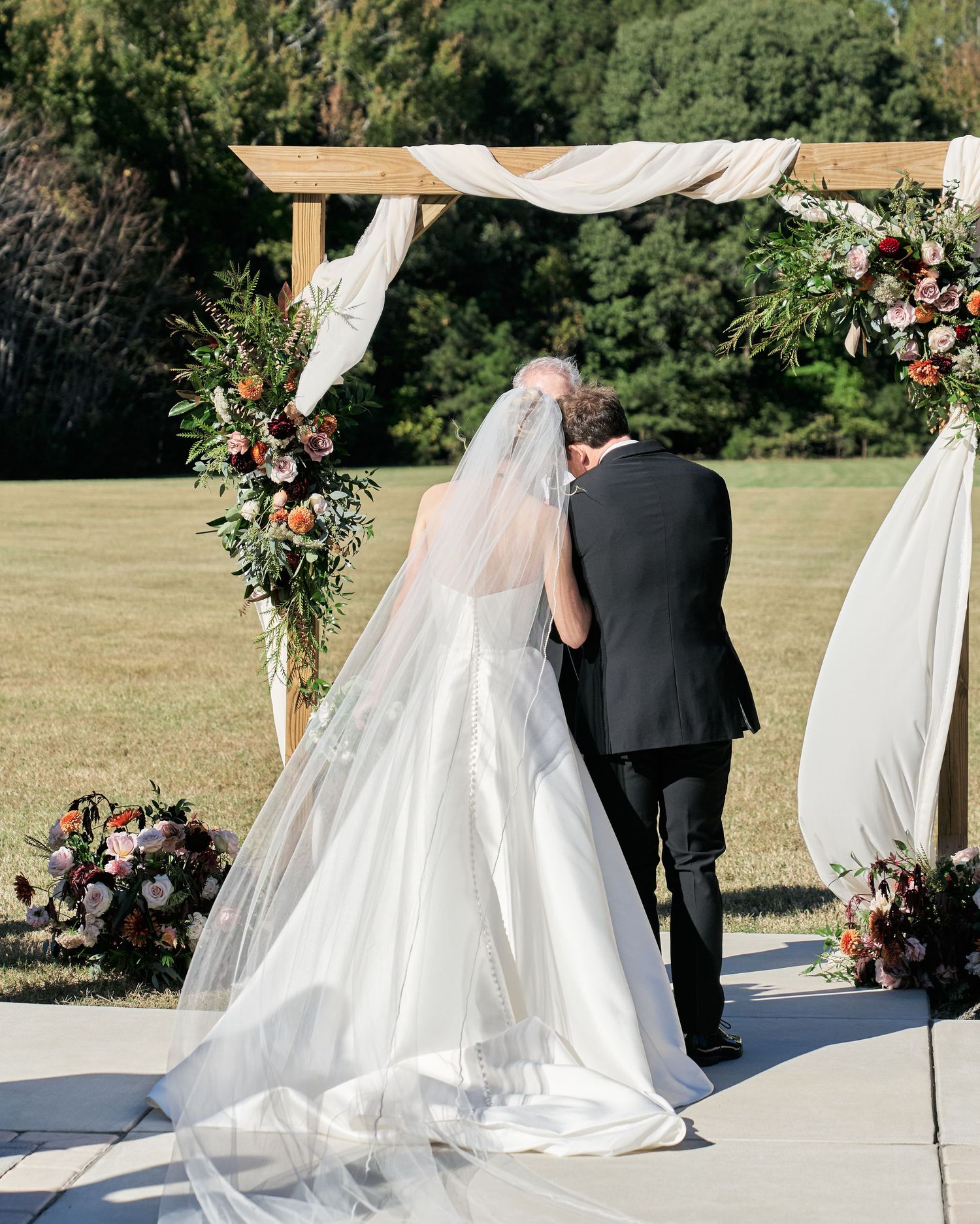 A wedding couple stands under a white canopy decorated with floral arrangements in an outdoor garden setting.