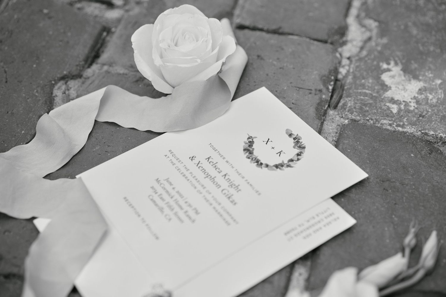 Black and white photo of wedding invitations with a white rose on stone pavement.