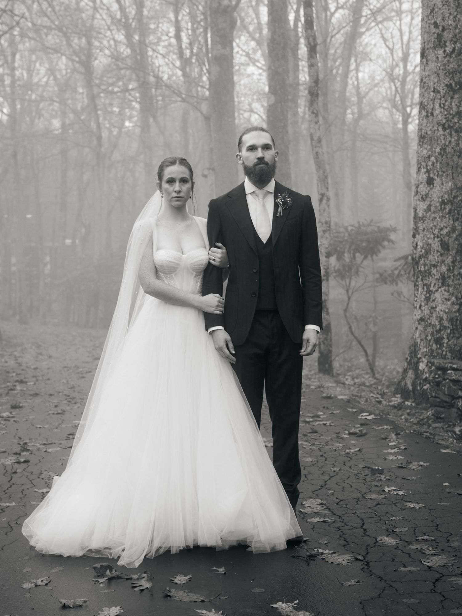 A moody black and white wedding portrait of a couple standing in a foggy forest setting.