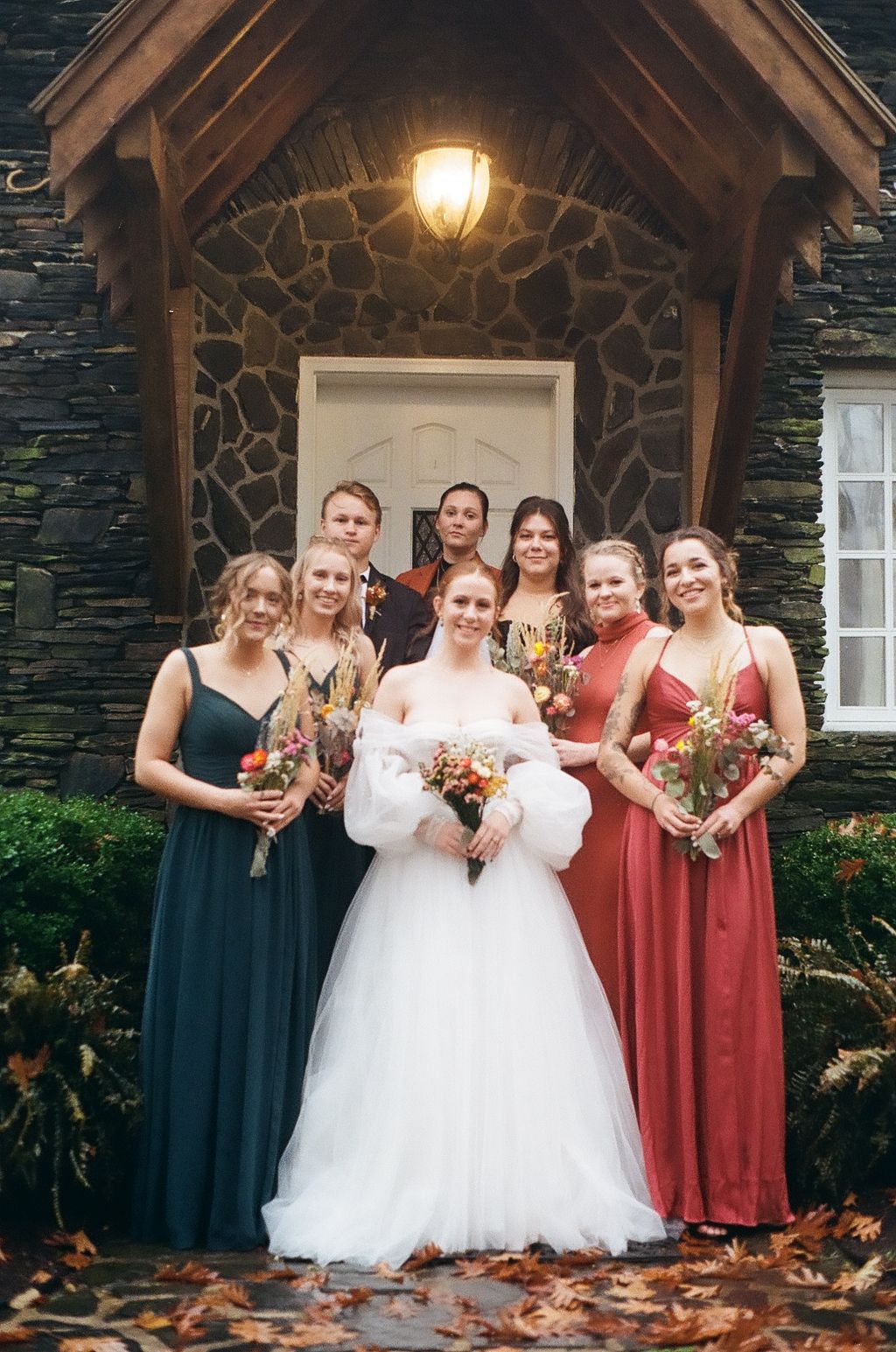 Bride in white gown poses with bridesmaids wearing teal and rust colored dresses under stone archway with glowing light.