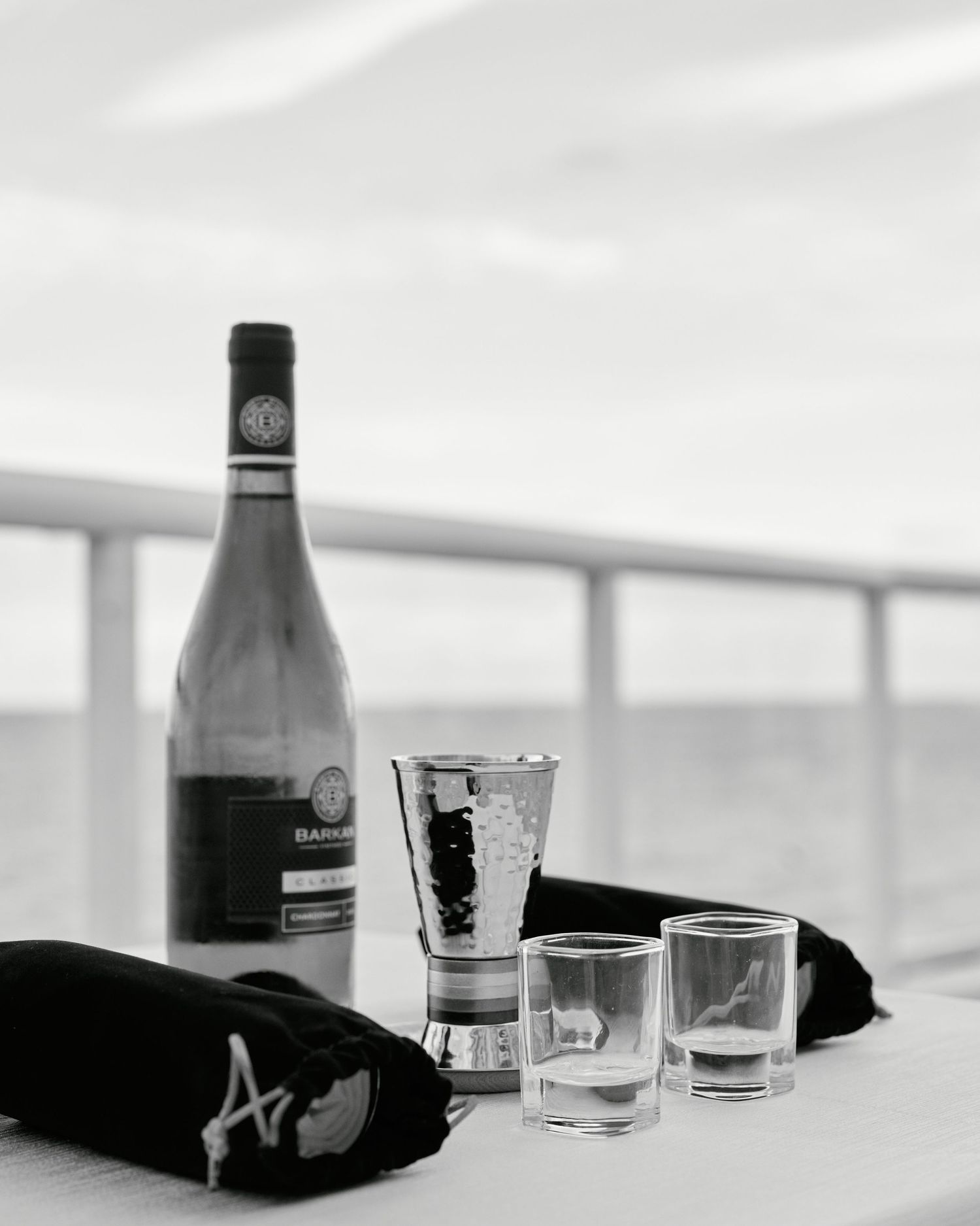 Black and white photo of wine bottle and glasses on outdoor balcony railing.