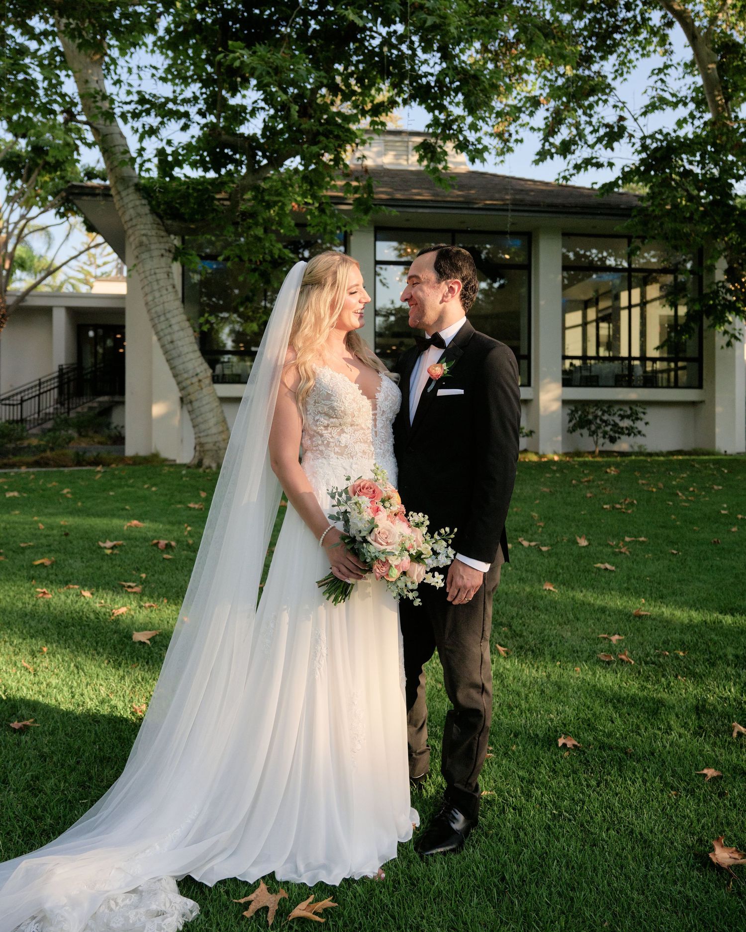 A couple poses for a wedding portrait on a grassy lawn with trees and architecture in the background.