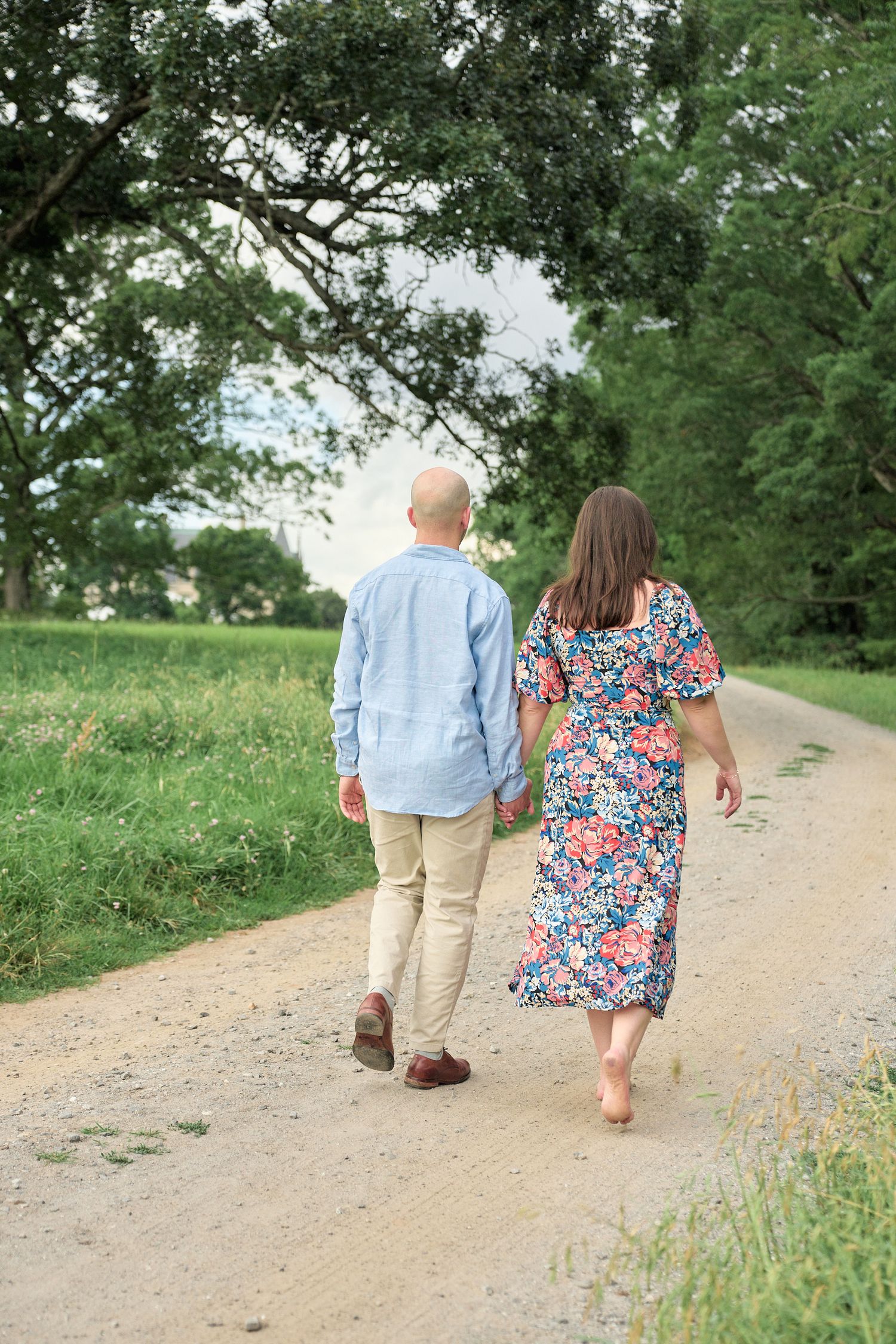 A couple walks hand in hand down a country path surrounded by green fields.