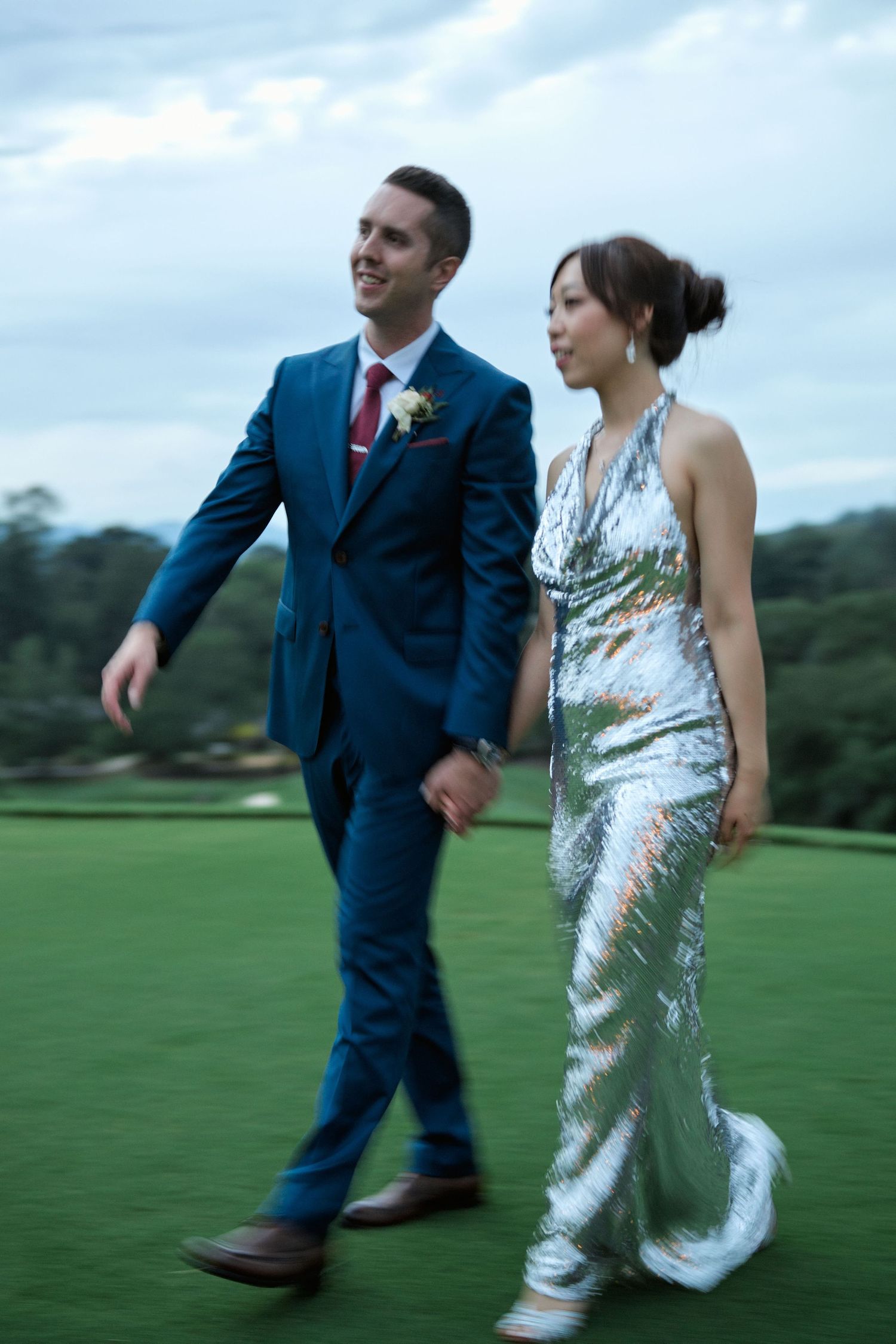 A couple taking a romantic stroll across a golf course in wedding attire during twilight.