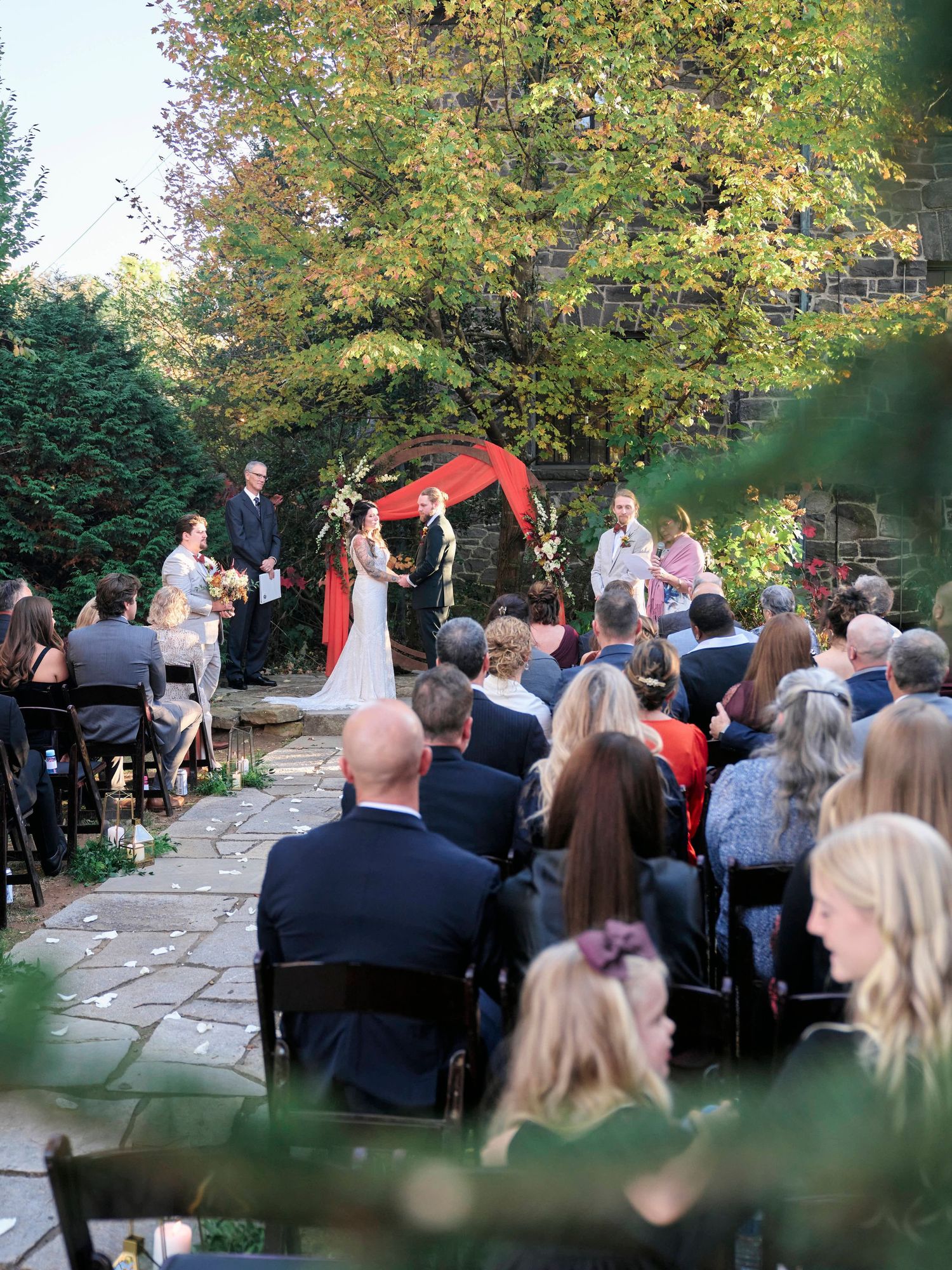 Outdoor wedding ceremony under fall foliage with guests seated on stone patio.