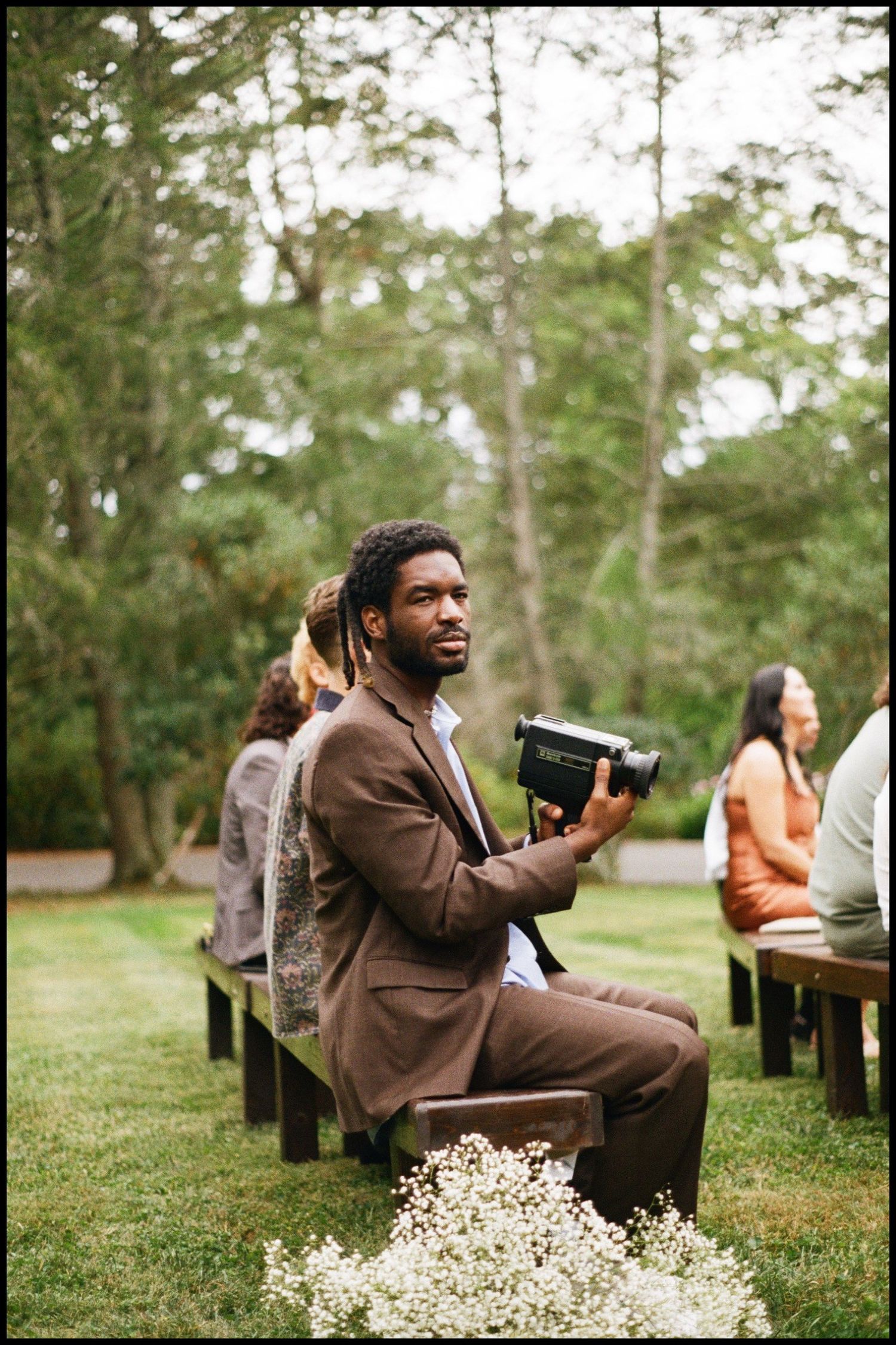 Wedding guest in brown suit holds camera while seated on wooden bench outdoors.