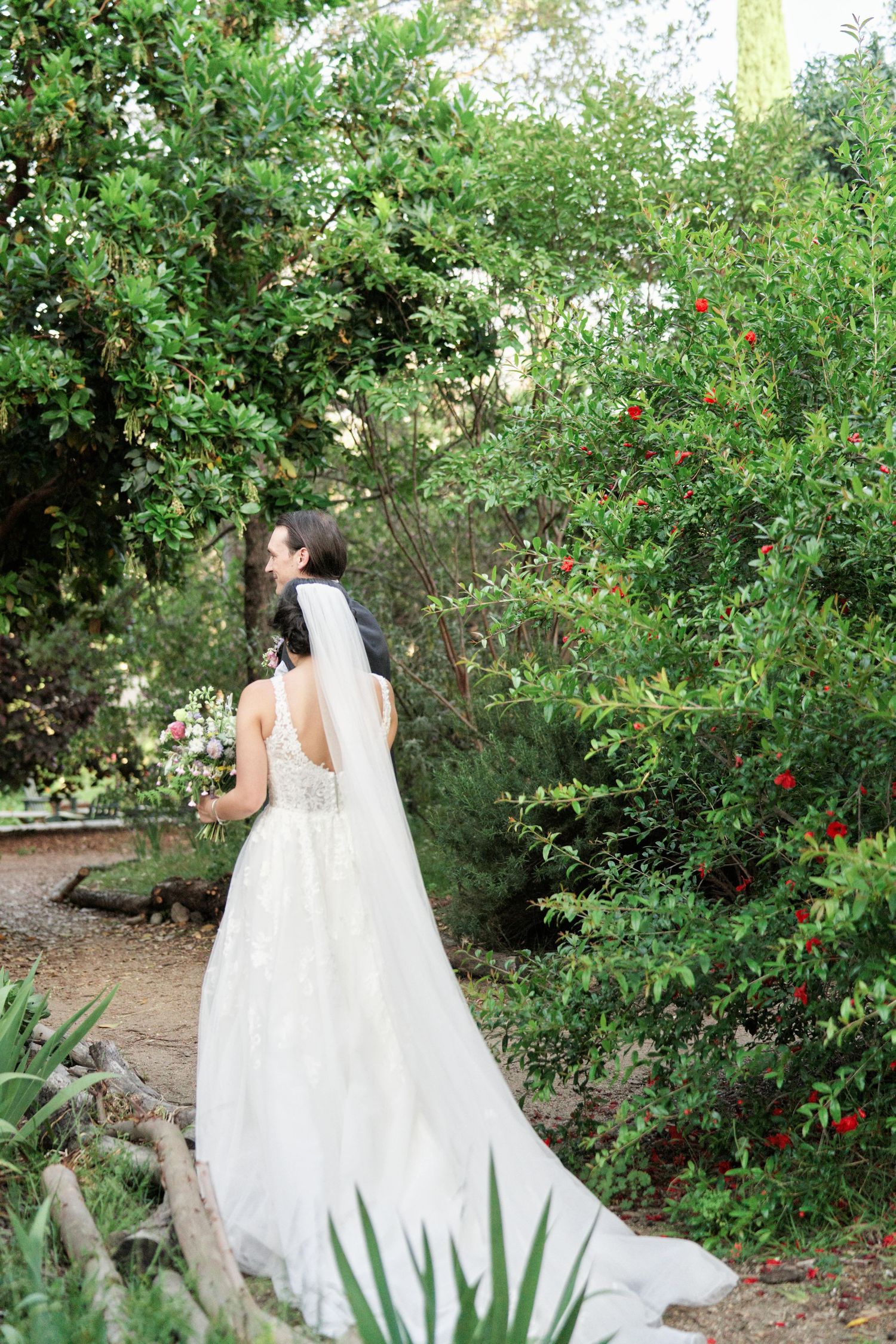 A bride in a flowing white gown poses in a lush garden setting with flowering bushes.