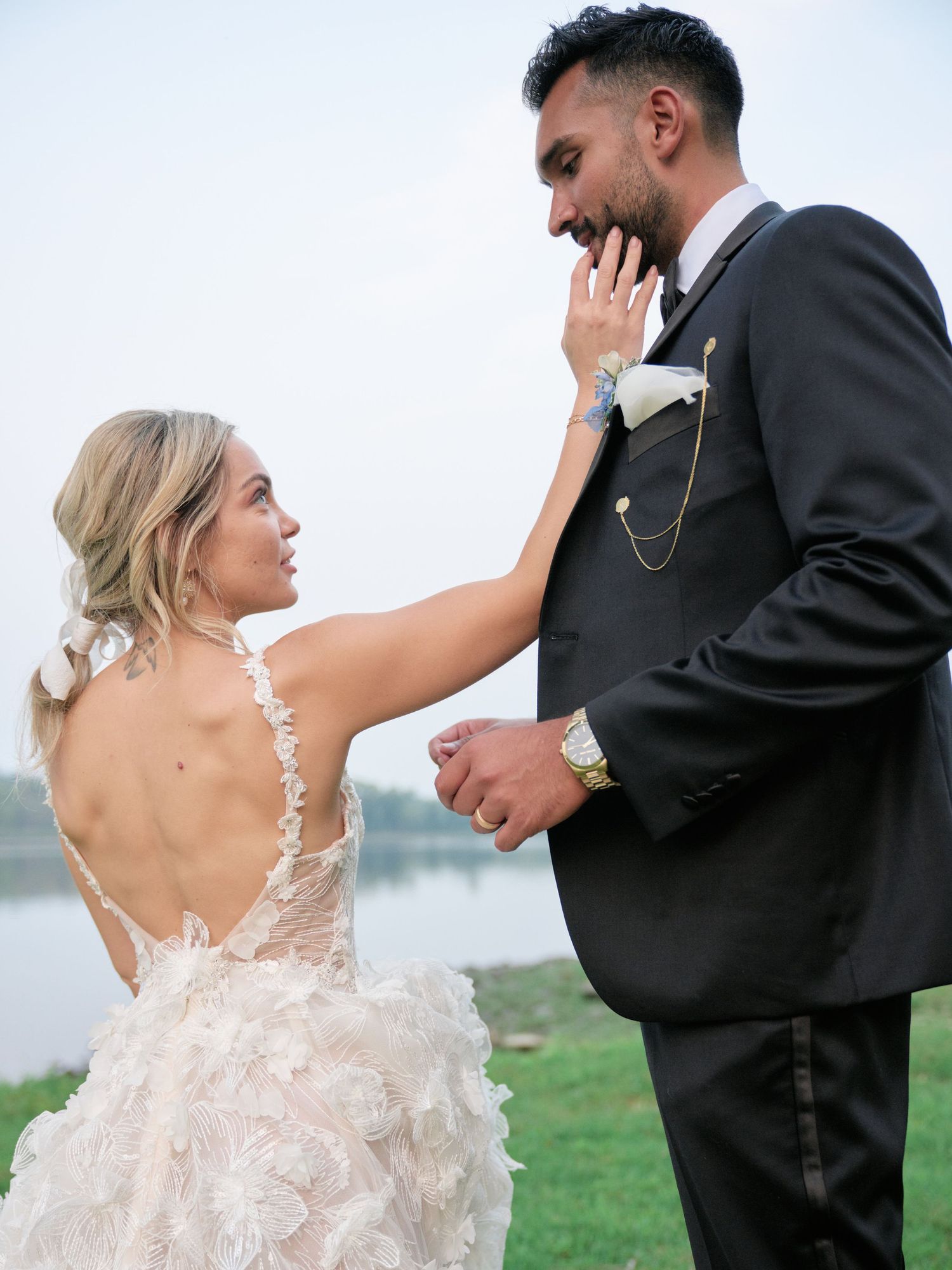 A couple shares an intimate moment on their wedding day wearing a floral lace gown and black tuxedo outdoors.