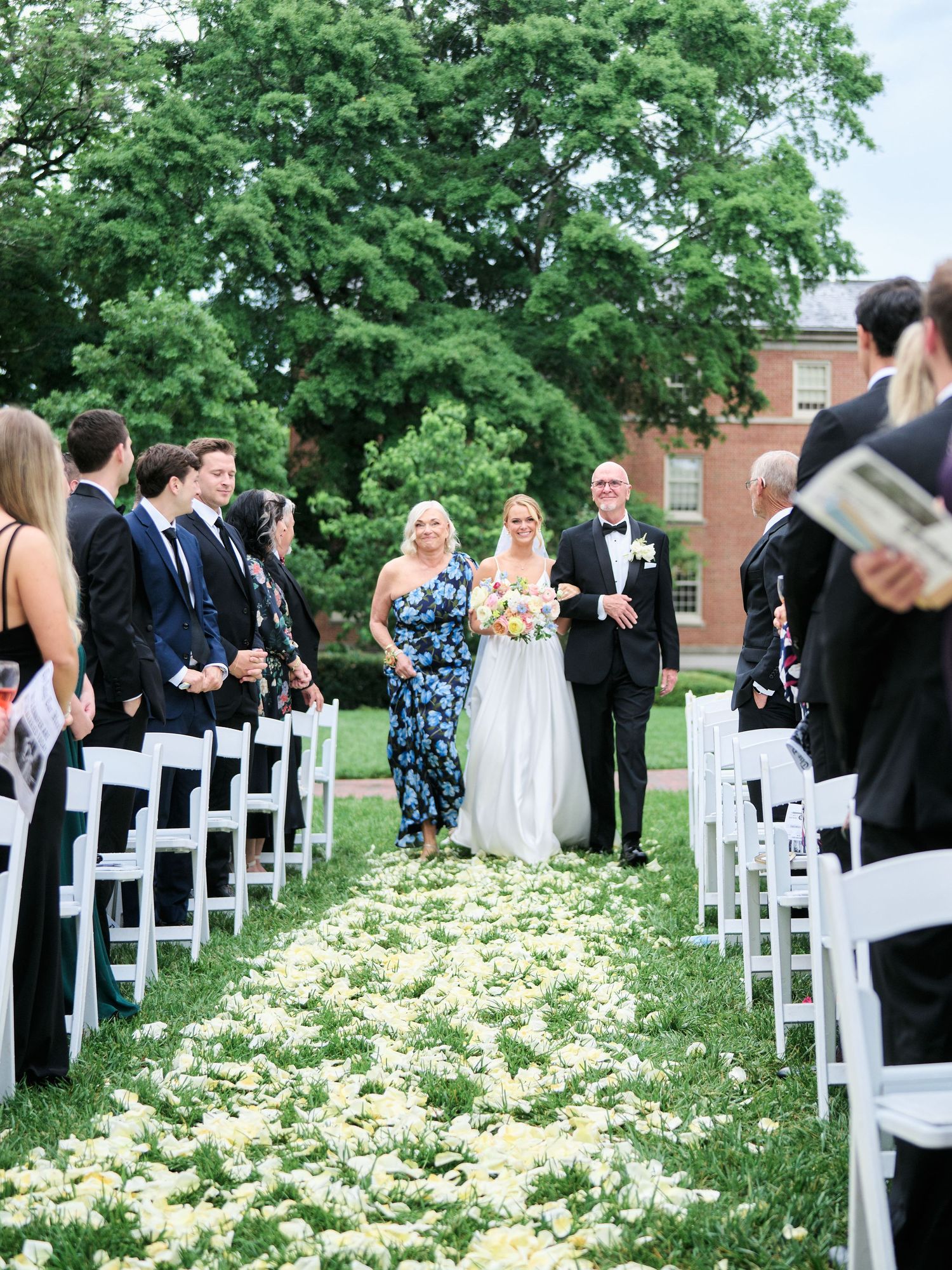 Wedding party walking down petal-covered aisle between white chairs during outdoor ceremony.
