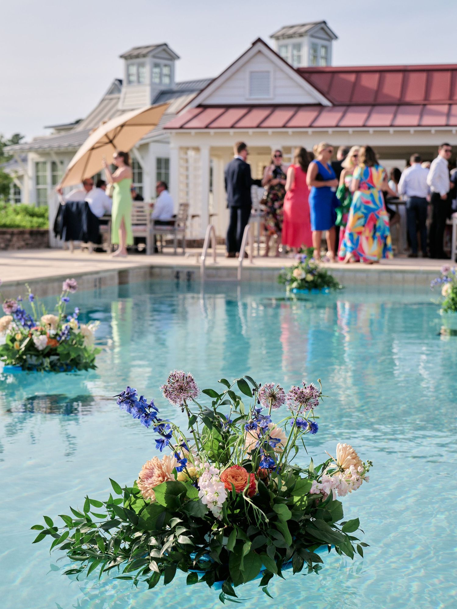Floral arrangement floats in pool at outdoor event with guests gathered under white tent near elegant house.