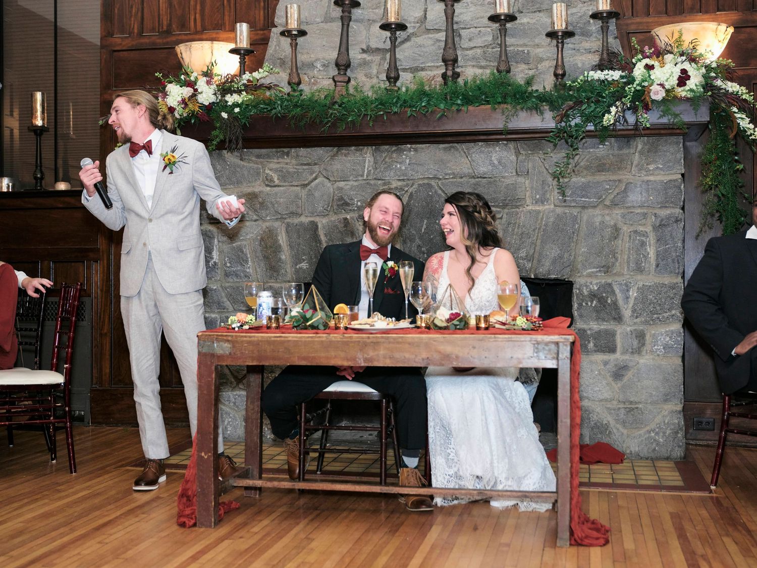 Wedding reception scene with couple at head table under stone fireplace and greenery garland decorations.