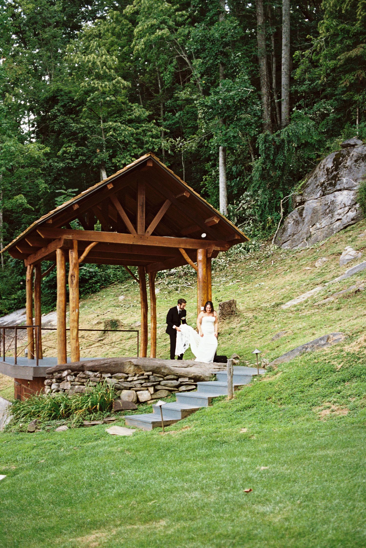 Bride and groom stand under a rustic wooden pavilion surrounded by lush forest and stone steps at an outdoor wedding venue.