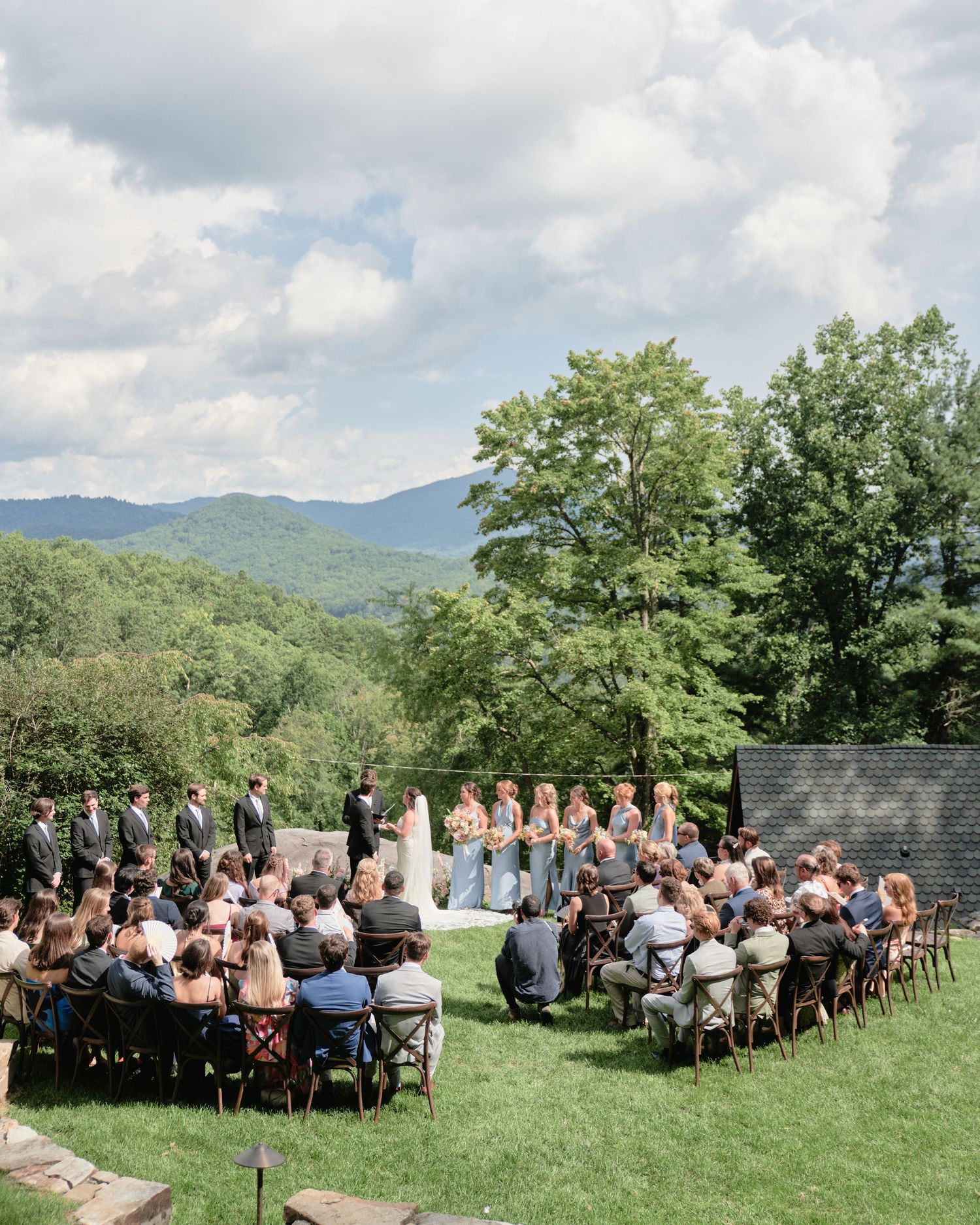 Outdoor wedding ceremony with guests seated on wooden chairs overlooking lush green mountains under a partly cloudy sky.