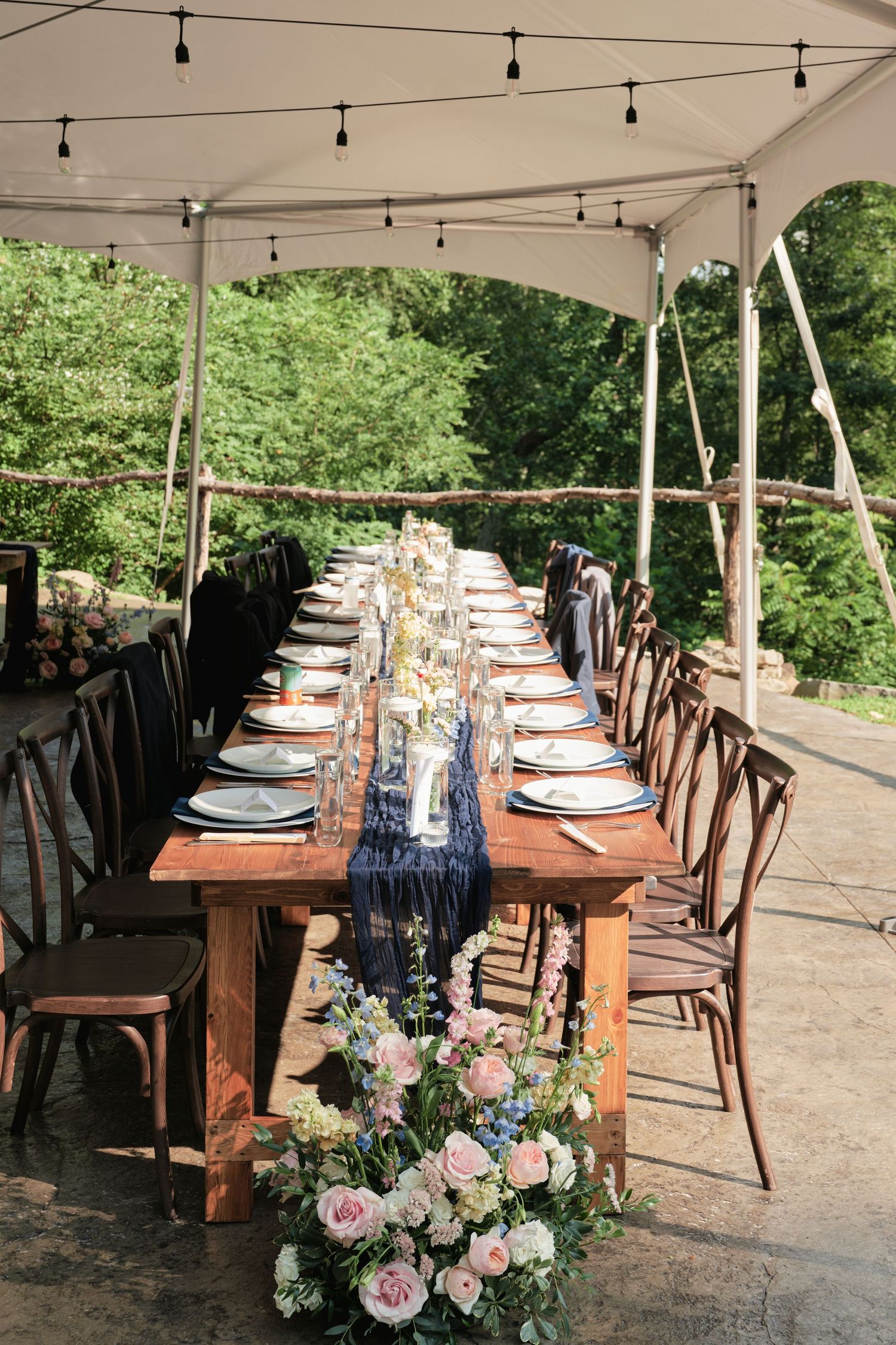 Rustic outdoor wedding reception table with navy runner, floral centerpieces, and string lights under a white tent.