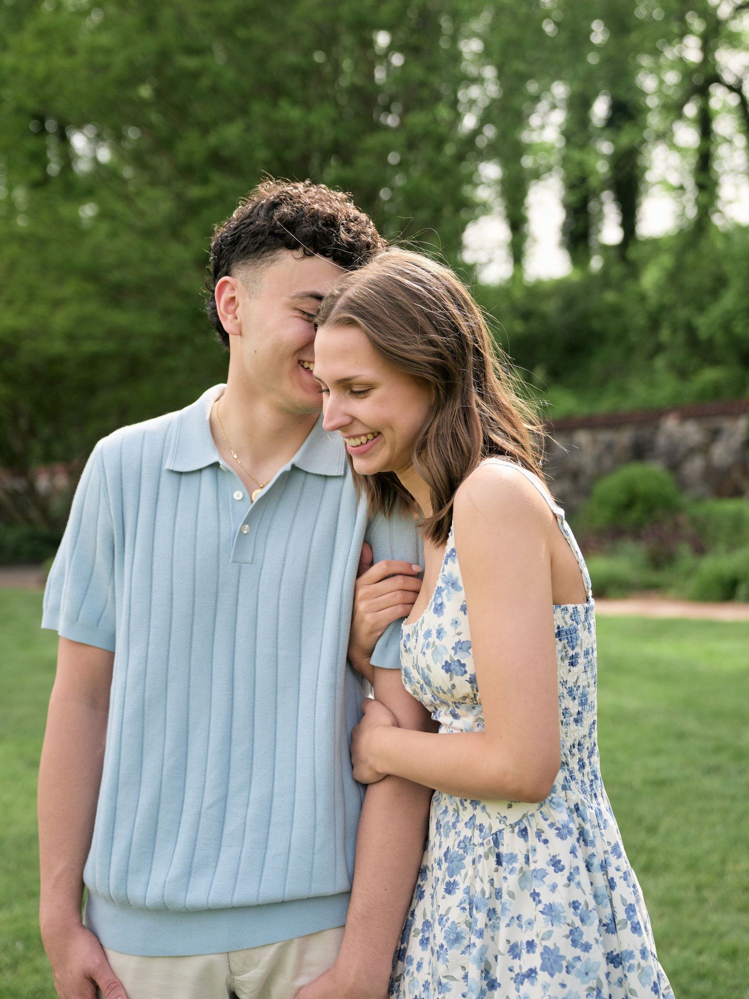 A young couple smiles and laughs together outdoors in a lush green garden, wearing light blue and floral outfits.