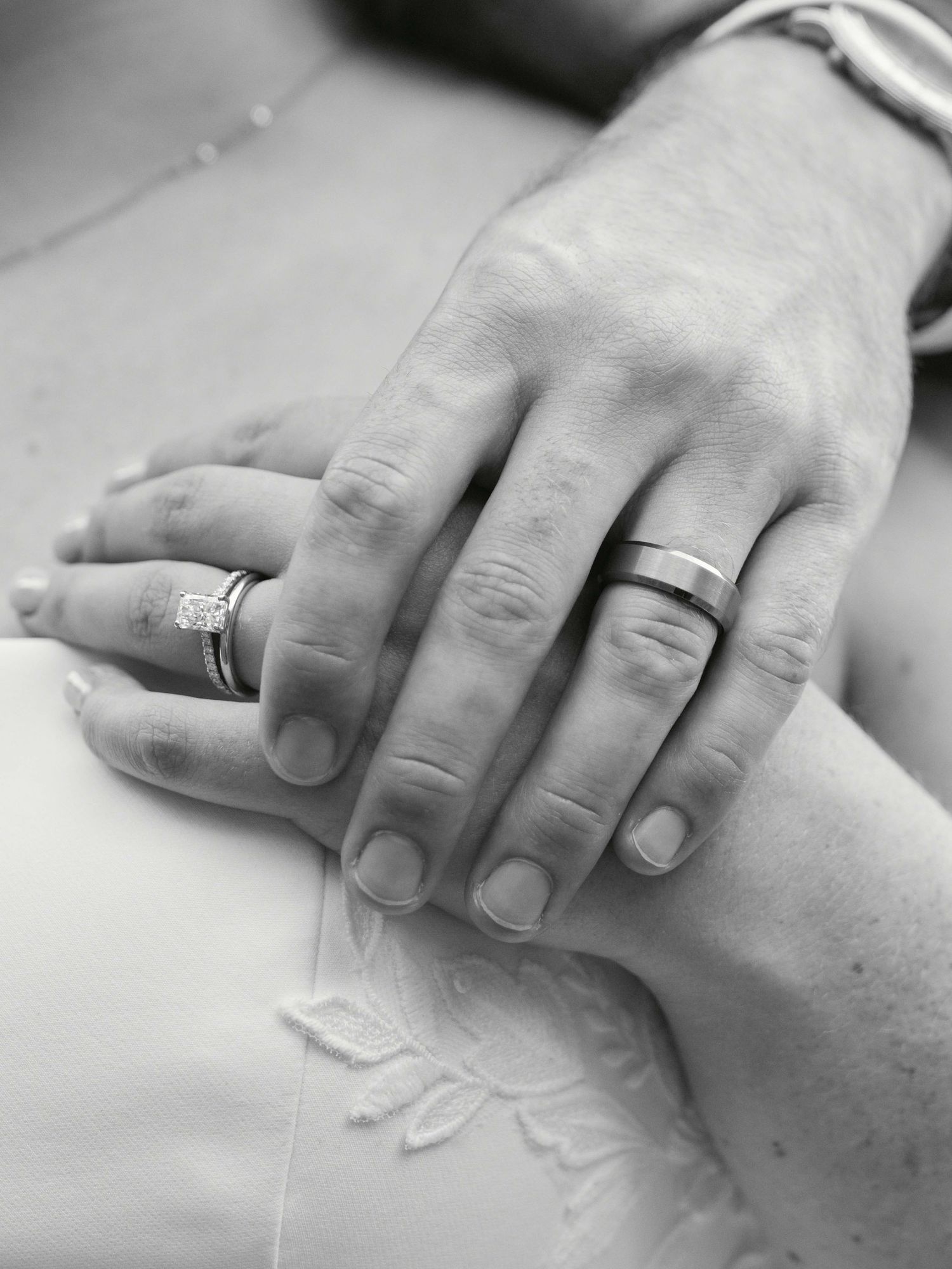 Black and white photo of two hands with wedding rings resting together on embroidered white fabric.