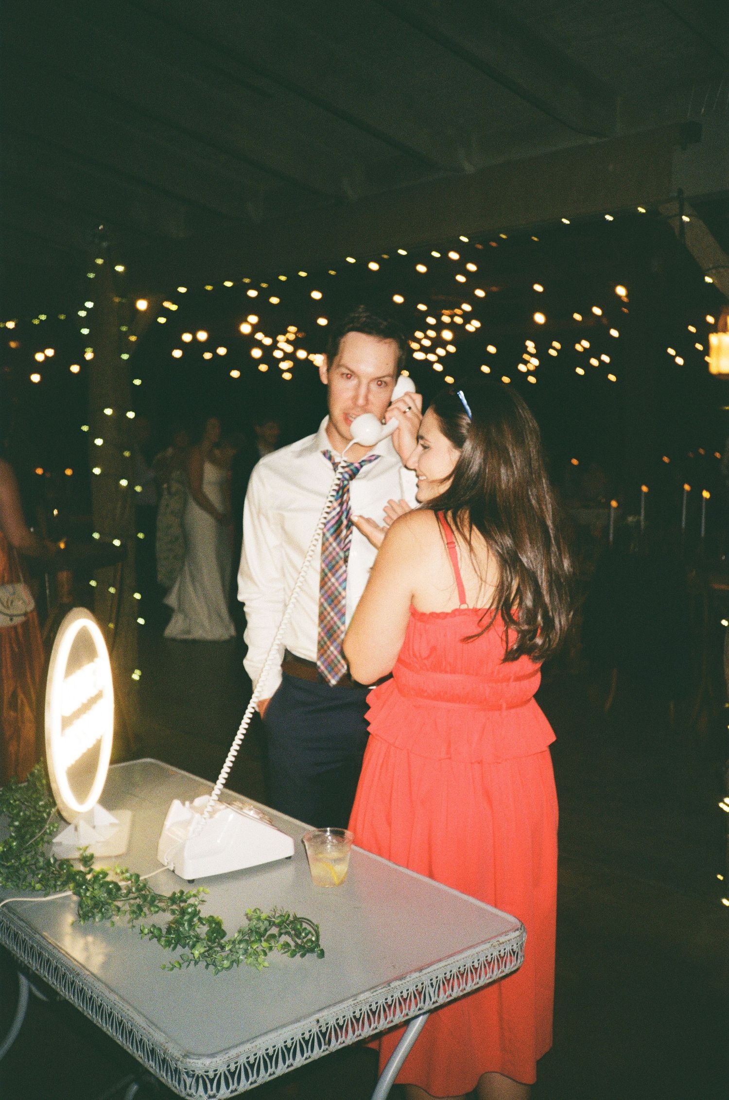 Couple sharing wedding cake at reception with twinkling lights backdrop, woman in red dress feeding groom in white shirt and tie.
