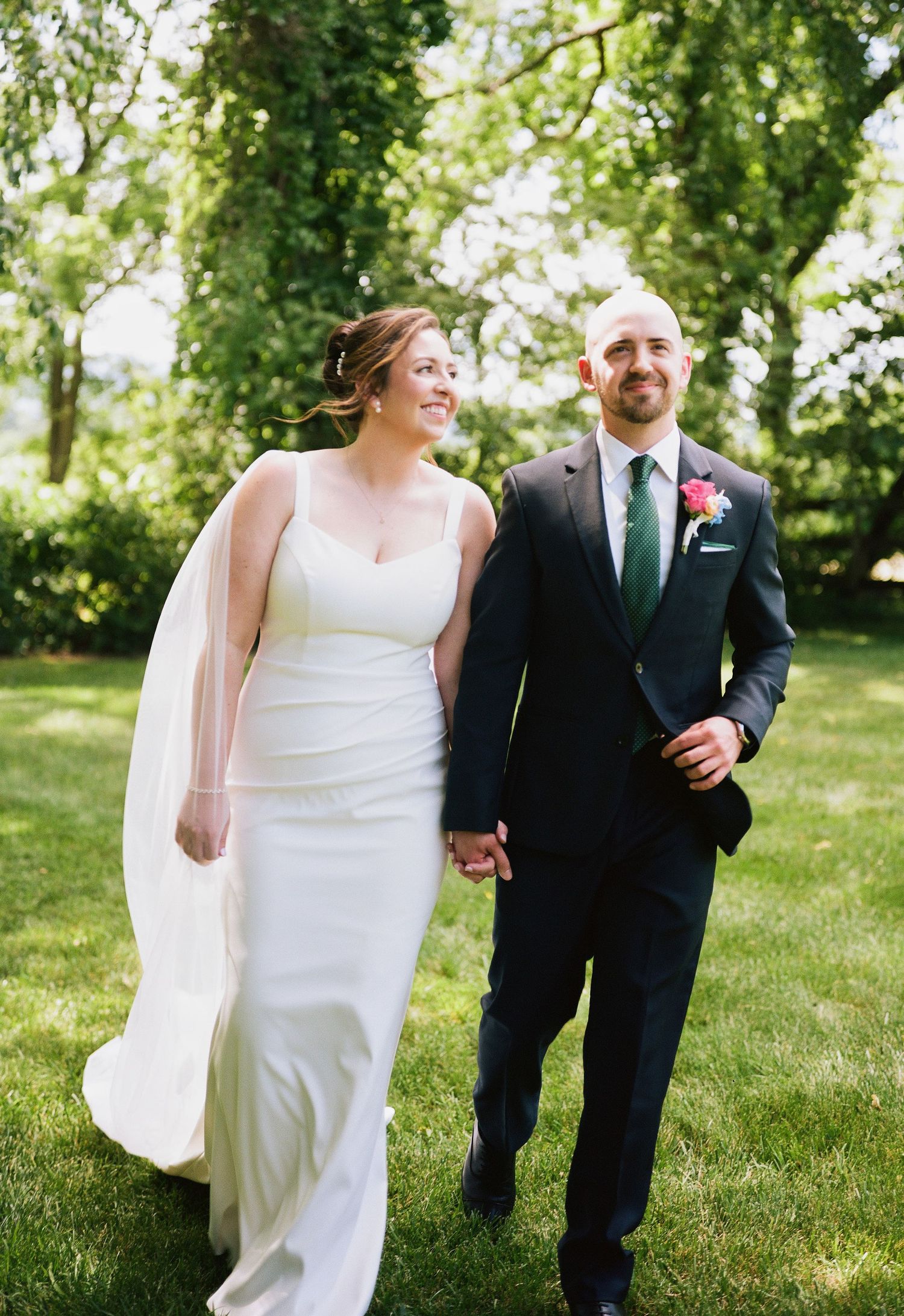 Bride in white gown and groom in navy suit holding hands, walking outdoors on green grass with trees behind them.