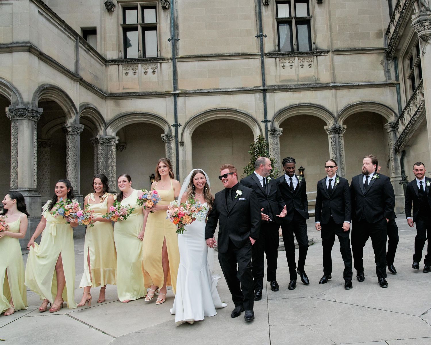 Wedding party in formal attire poses in a line against a stone building with arched columns and architectural details.
